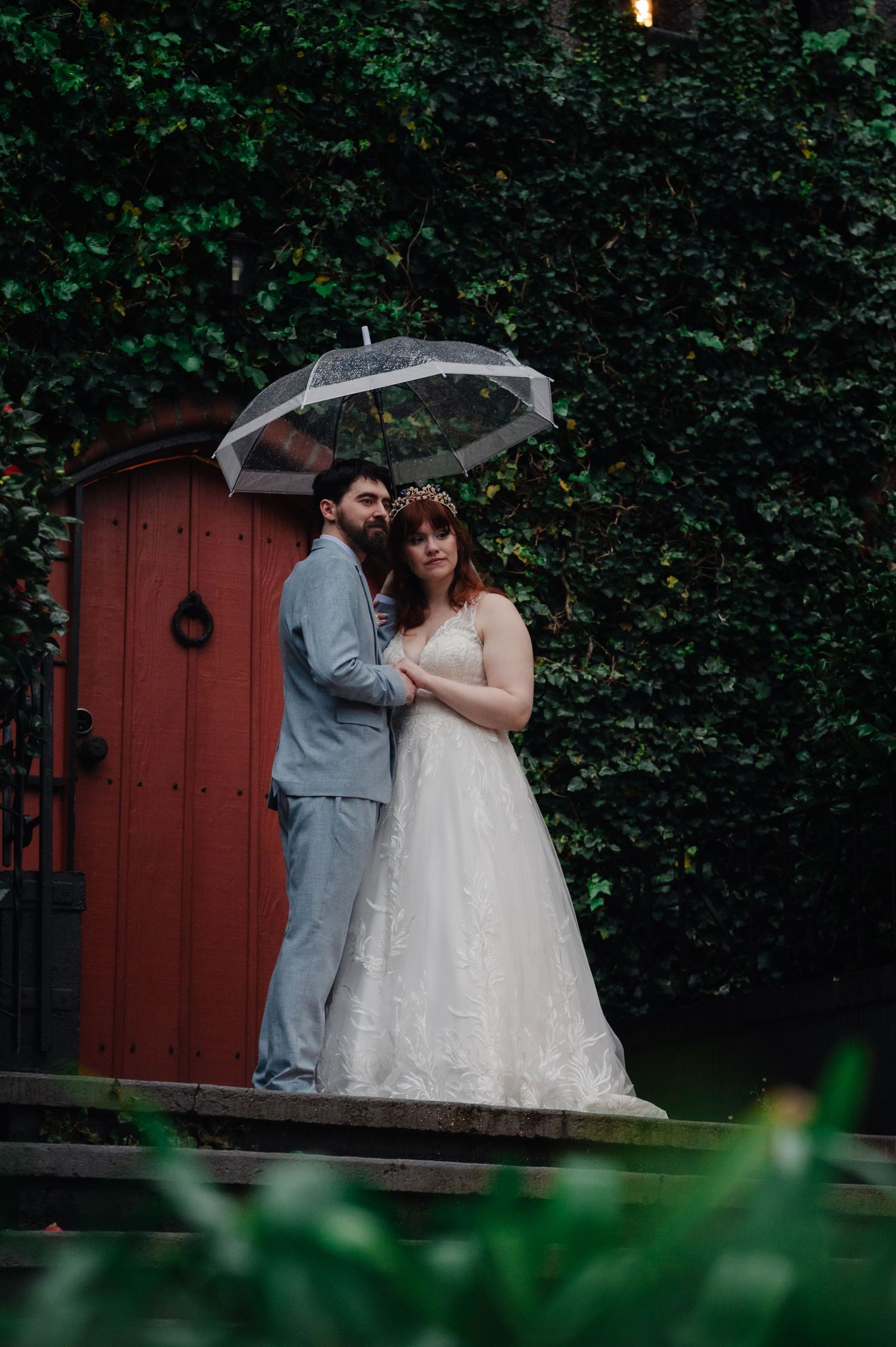 Wedding couple shares intimate moment in front of red garden door surrounded by lush greenery.