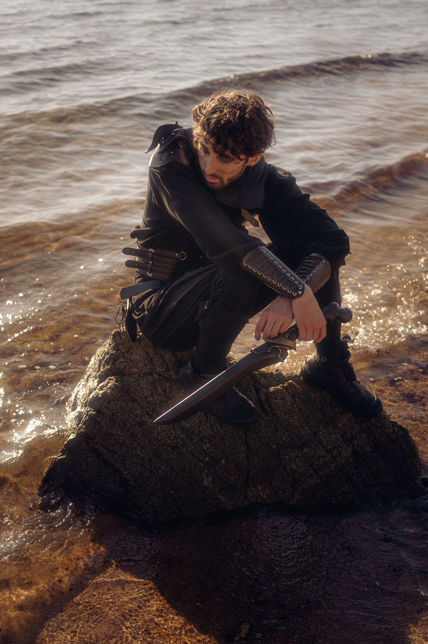 Silhouette figure crouches on rock by water at sunset holding dagger.