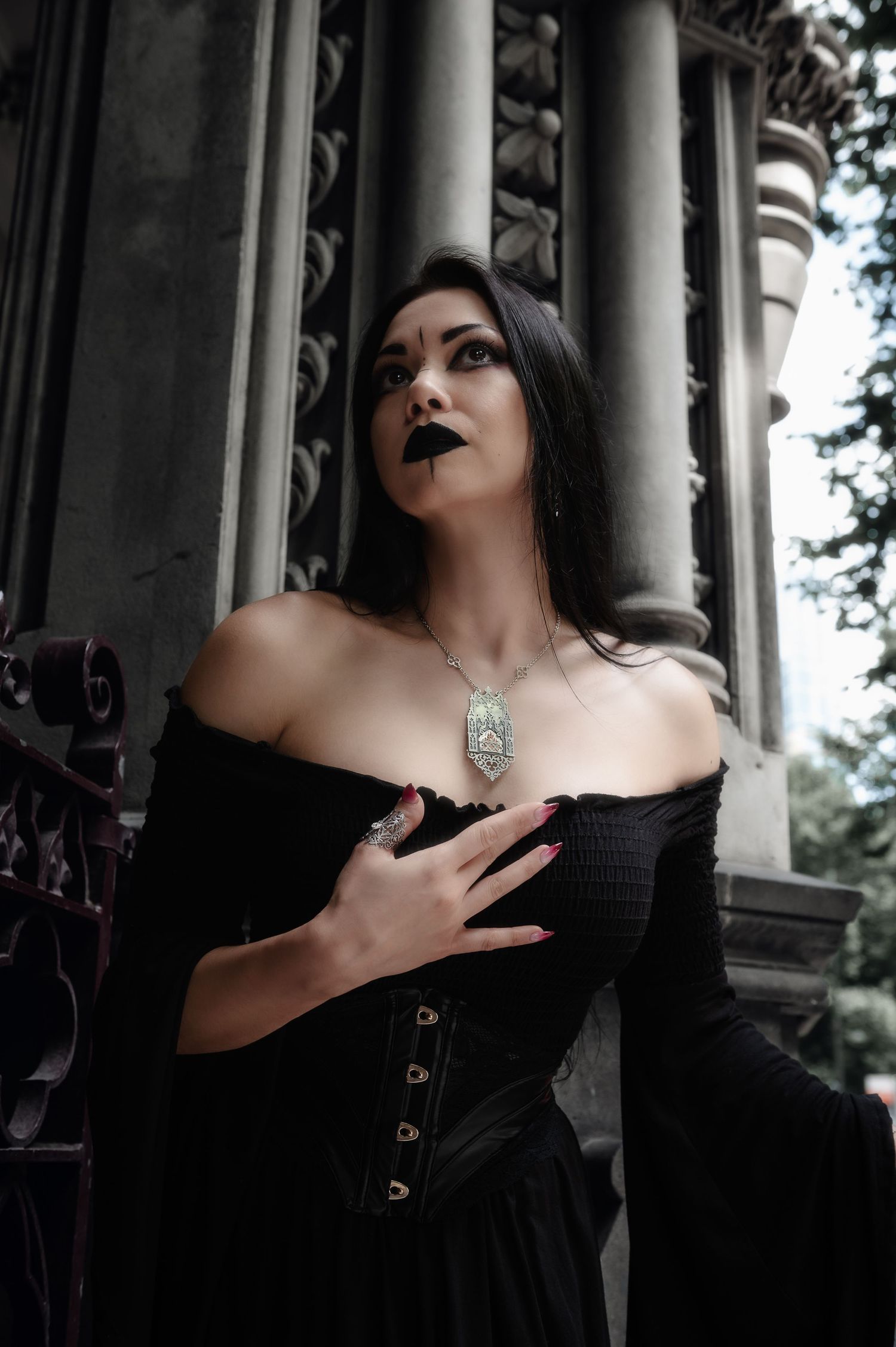Gothic-inspired portrait in black dress with ornate silver necklace against classical stone columns.