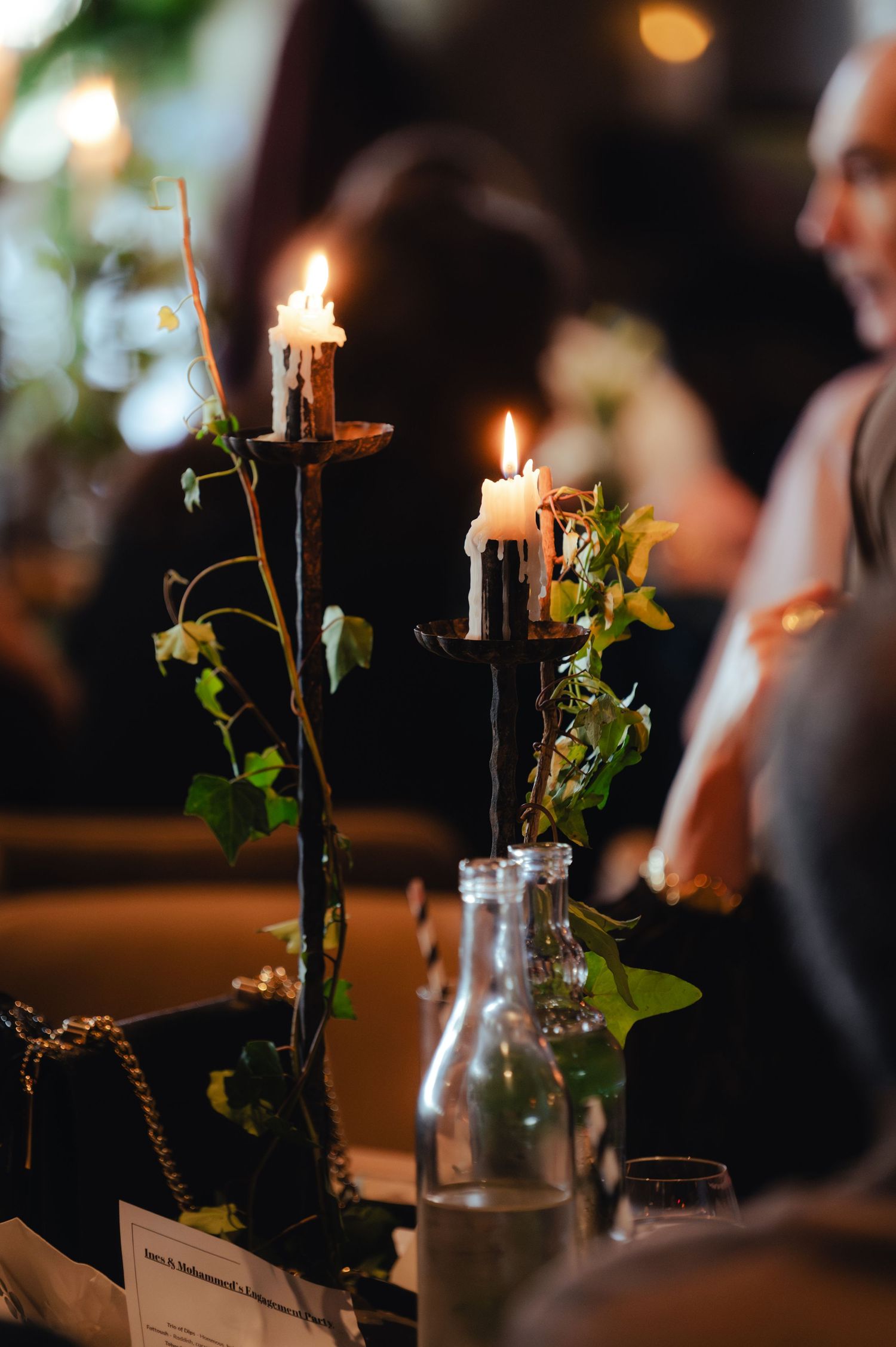 Elegant candlelit table centerpiece with twisted black candle holders, ivy vines, and glass bottles at a romantic evening event.