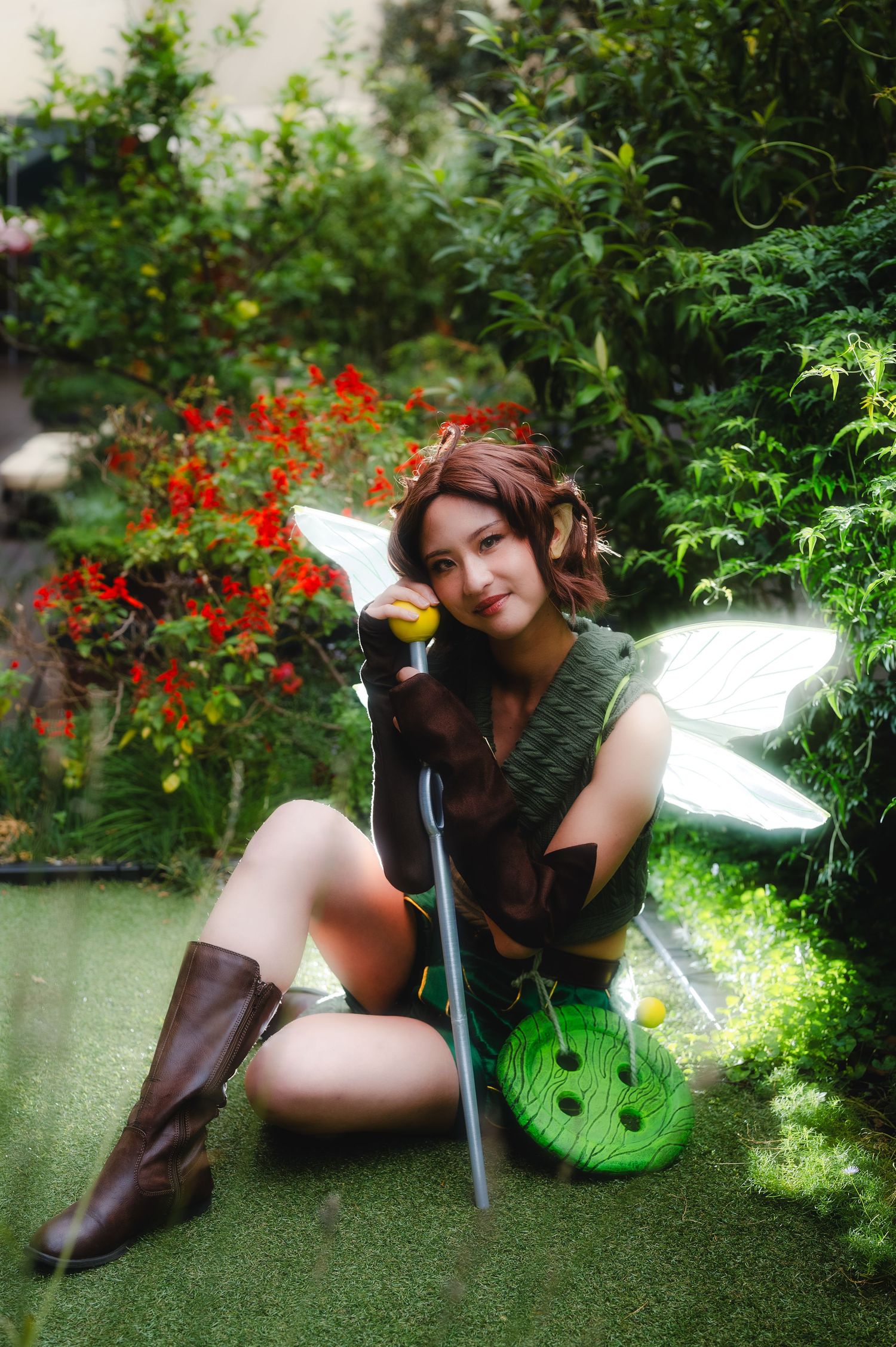 A woman cosplaying as a fairy character in a green outfit with wings, brown boots, and a staff, posing in a lush garden.