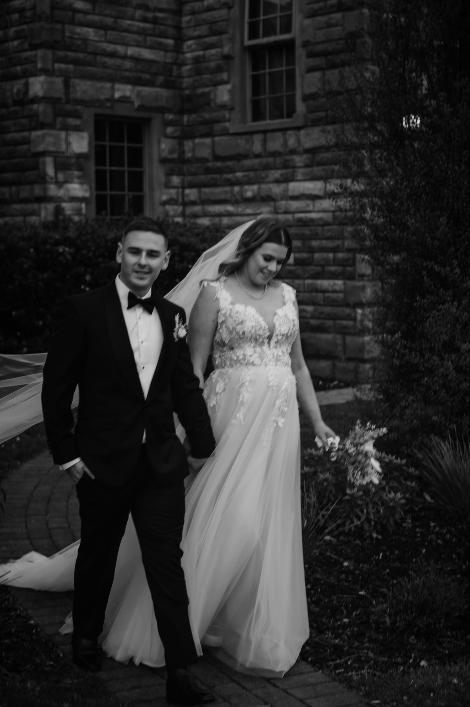 Wedding couple walking together outside a stone building in formal attire.