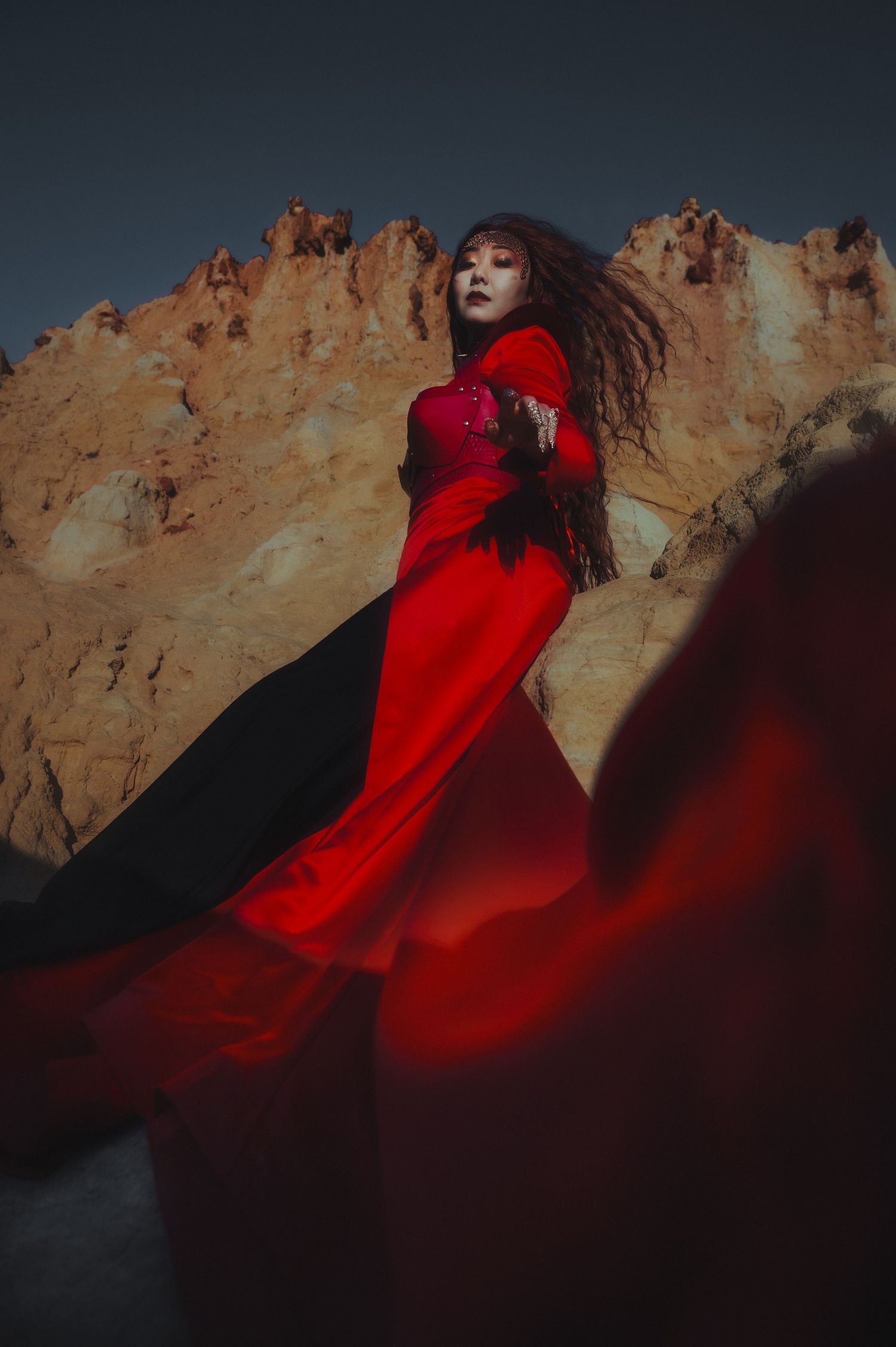 A dramatic red gown billows against rocky cliffs in moody lighting.
