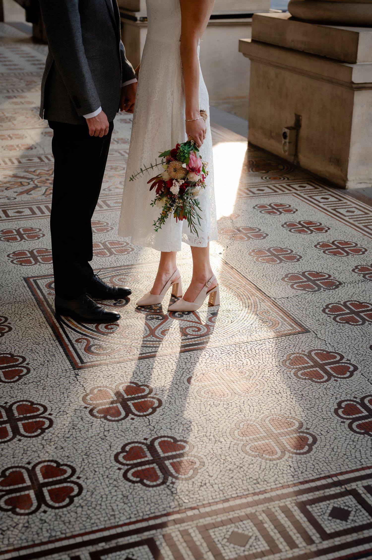 Wedding shoes and dress hem on ornate patterned tile floor with dramatic lighting.