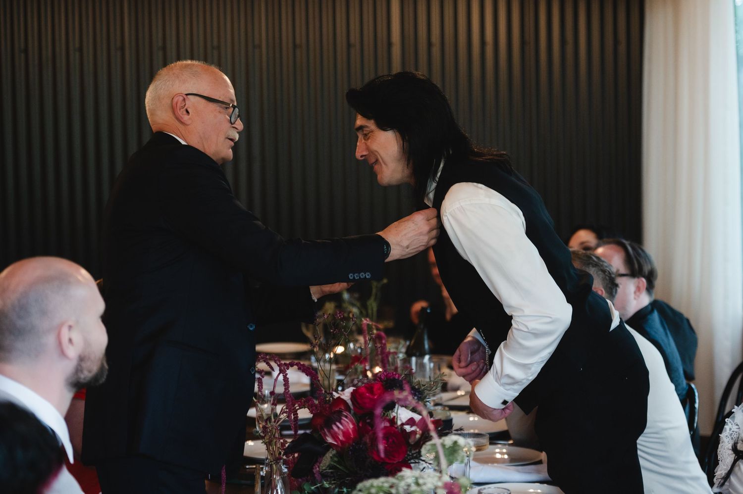 Two men share a warm moment at an elegant wedding reception dinner, one adjusting the other's tie.
