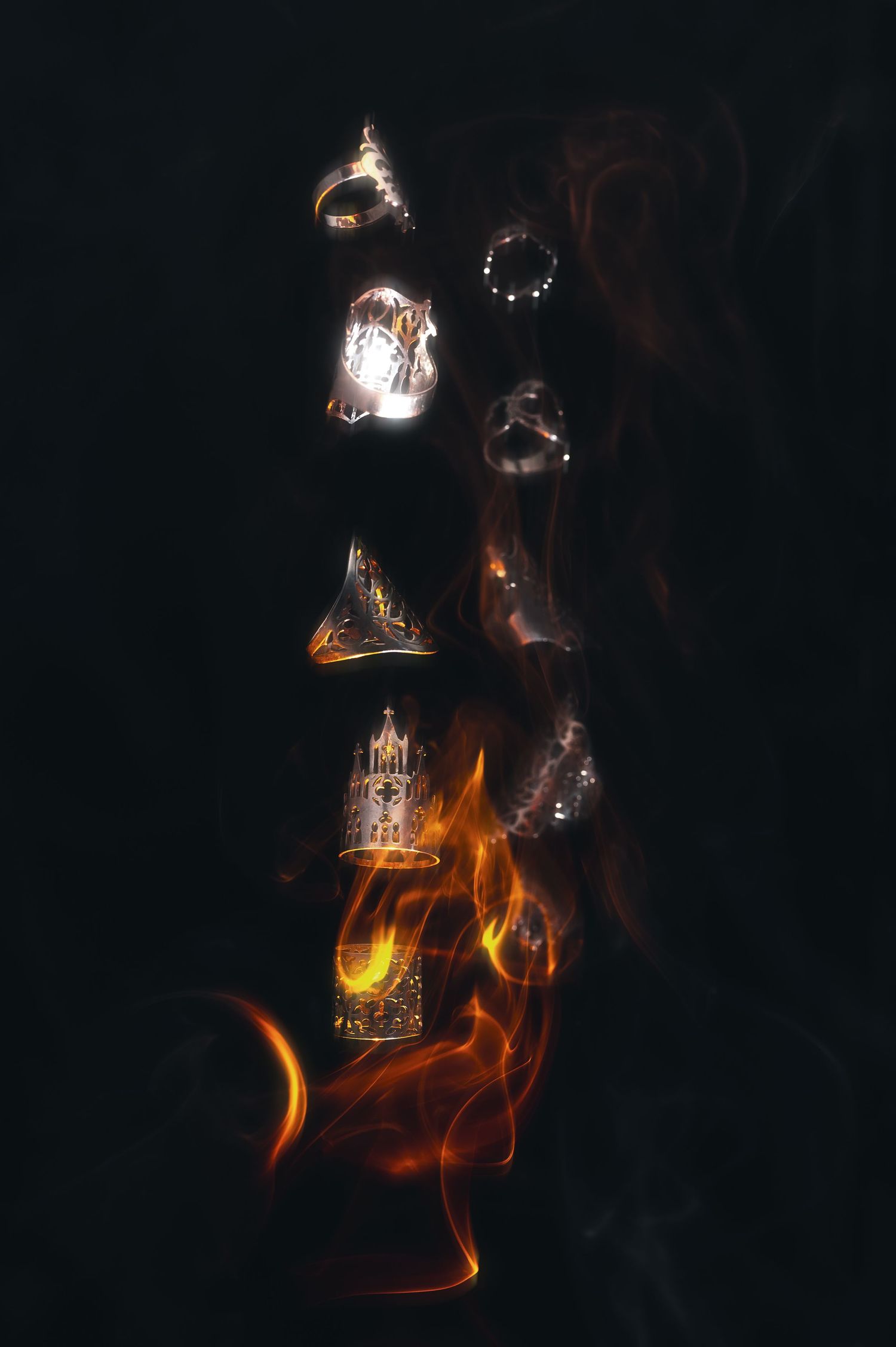 Dark mystical arrangement of ornate rings and jewelry floating in smoke above dramatic orange flames on black background.