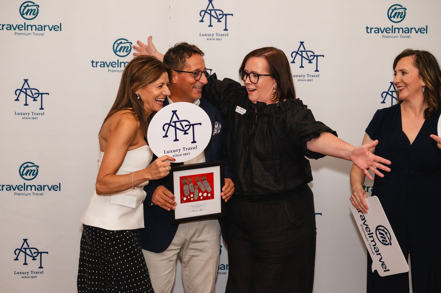 A group of people pose and laugh together in front of a branded step-and-repeat backdrop at a formal event.