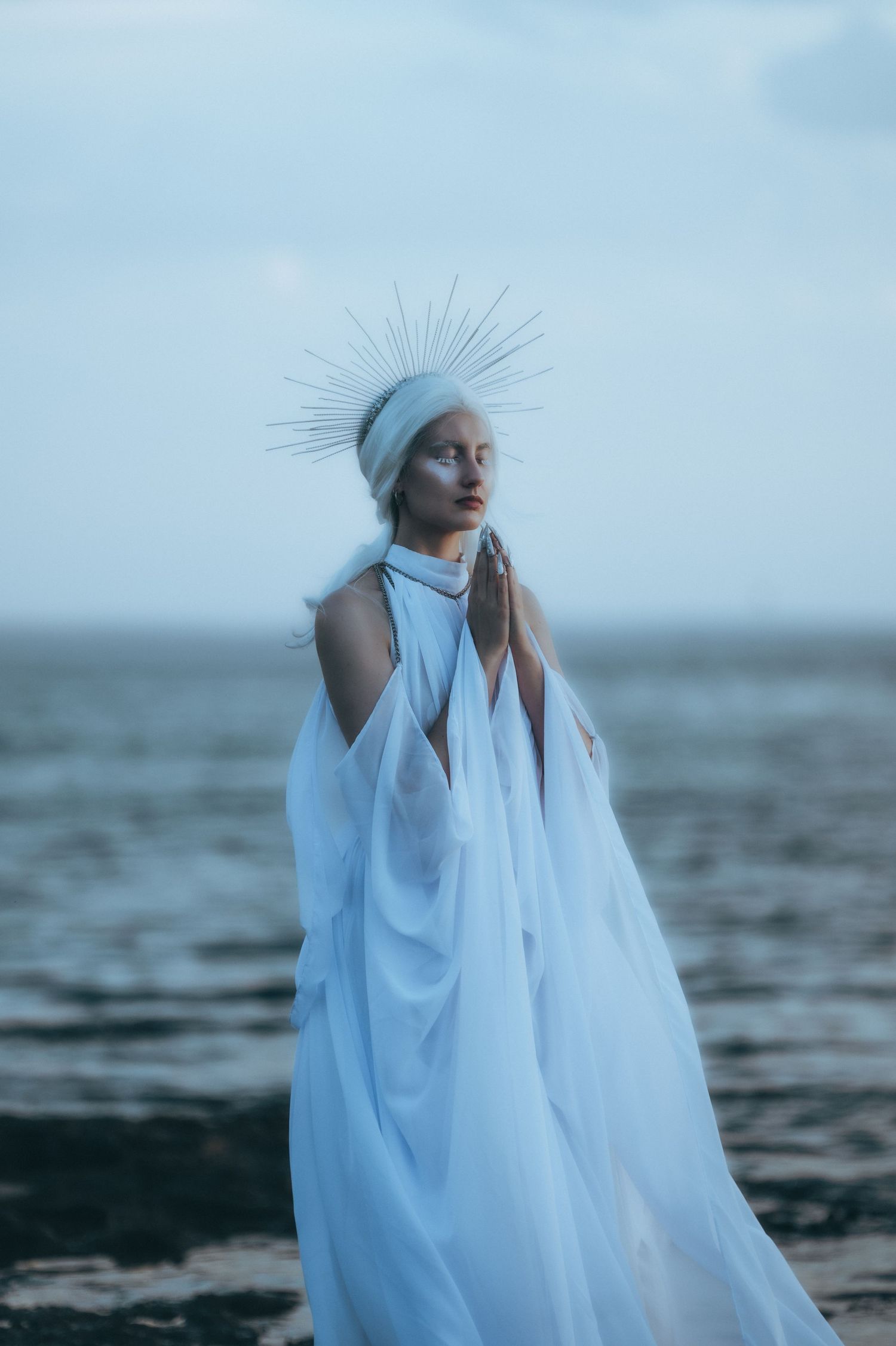 Ethereal figure in flowing white fabric and blue headpiece poses against moody ocean backdrop at dusk.
