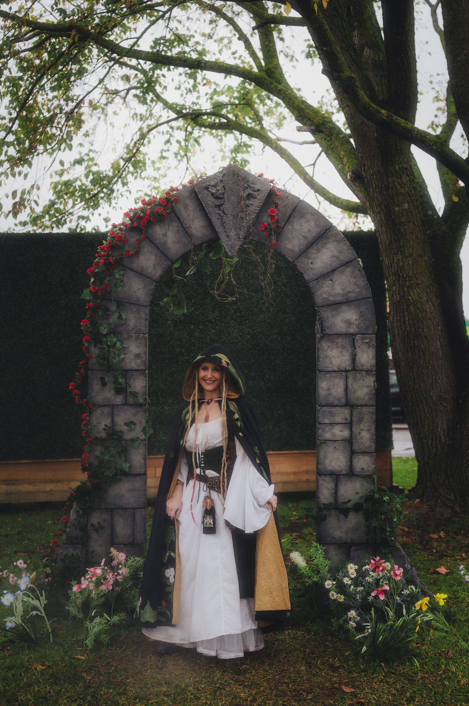 Woman in medieval fantasy costume stands under a stone arch decorated with flowers and ivy in a garden setting.