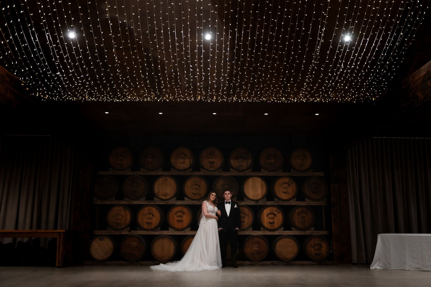 Wedding venue decorated with twinkling fairy lights and wine barrels along the wall.