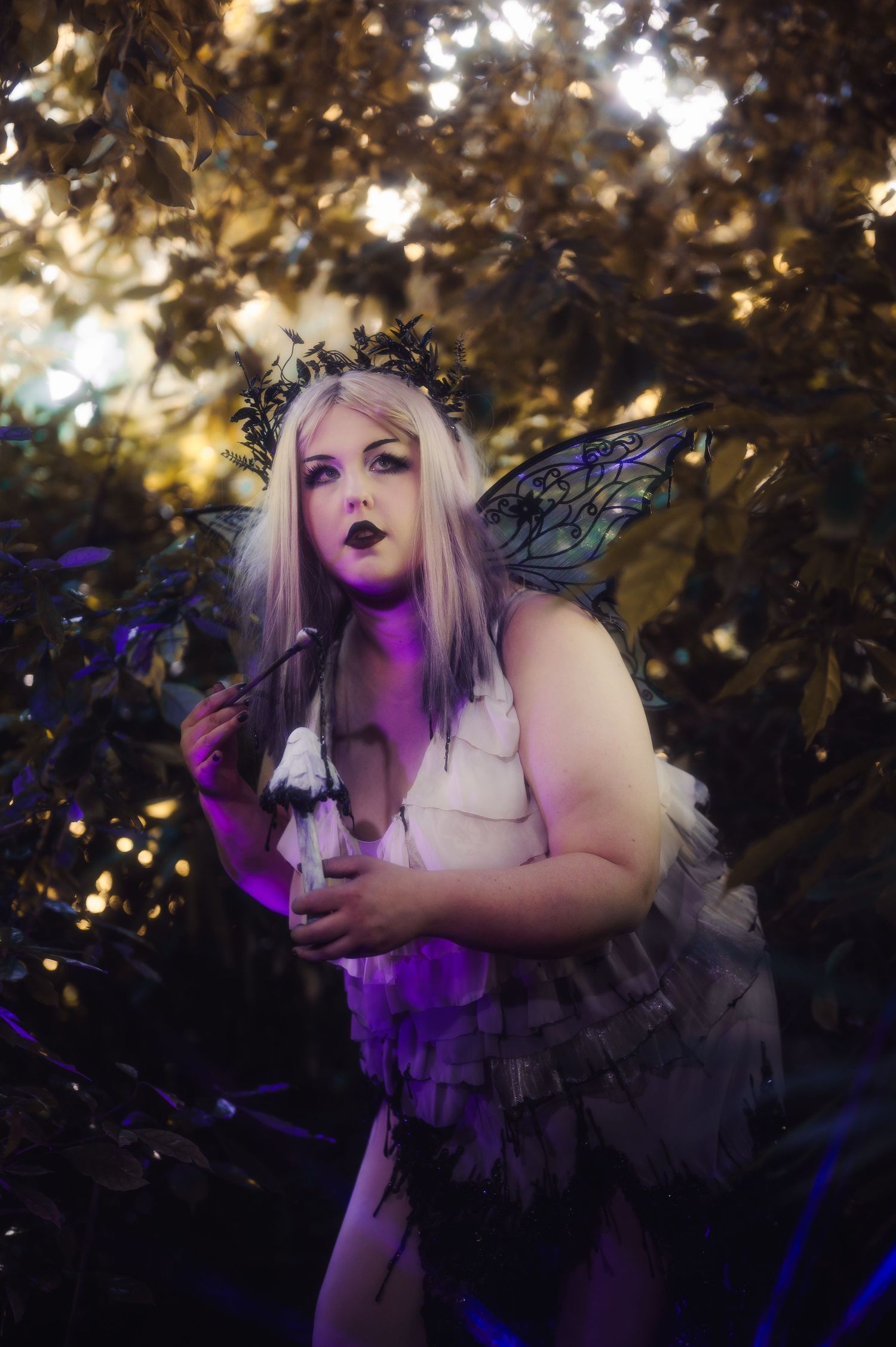 A woman in a dark fairy costume with wings and floral crown poses dramatically among lush forest foliage in purple light.