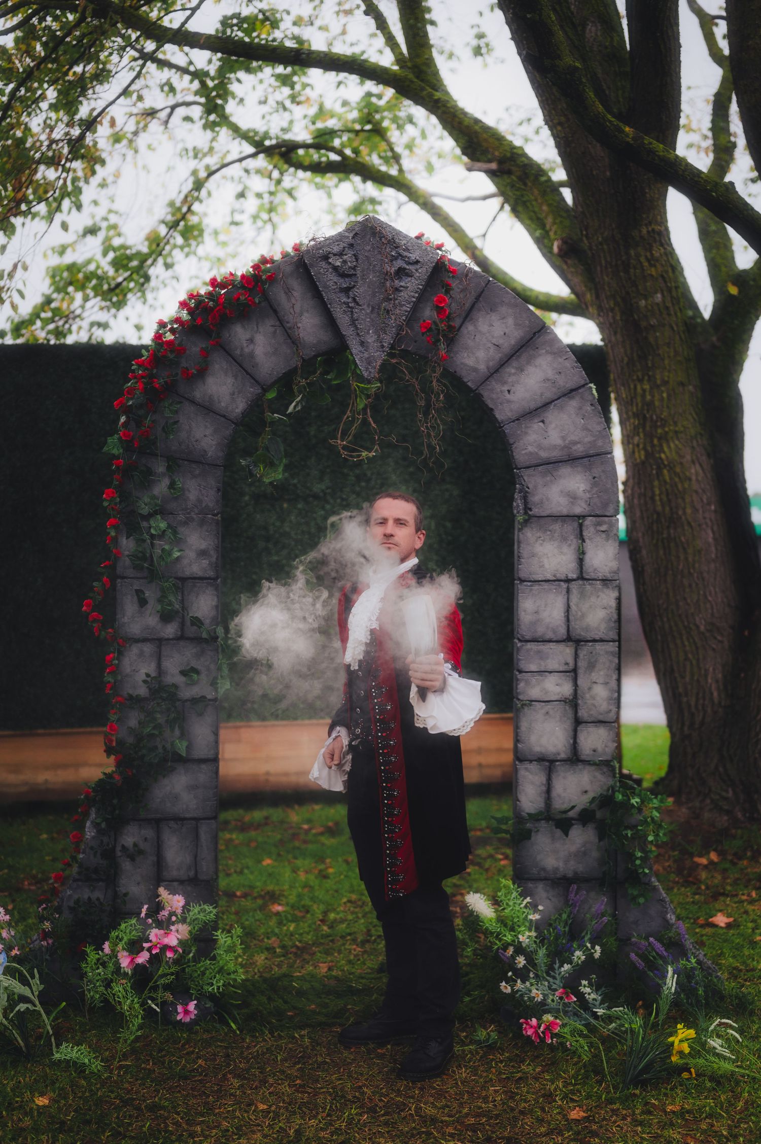 Man in Victorian costume stands before a stone arch with red flowers and smoke effects in a garden setting.