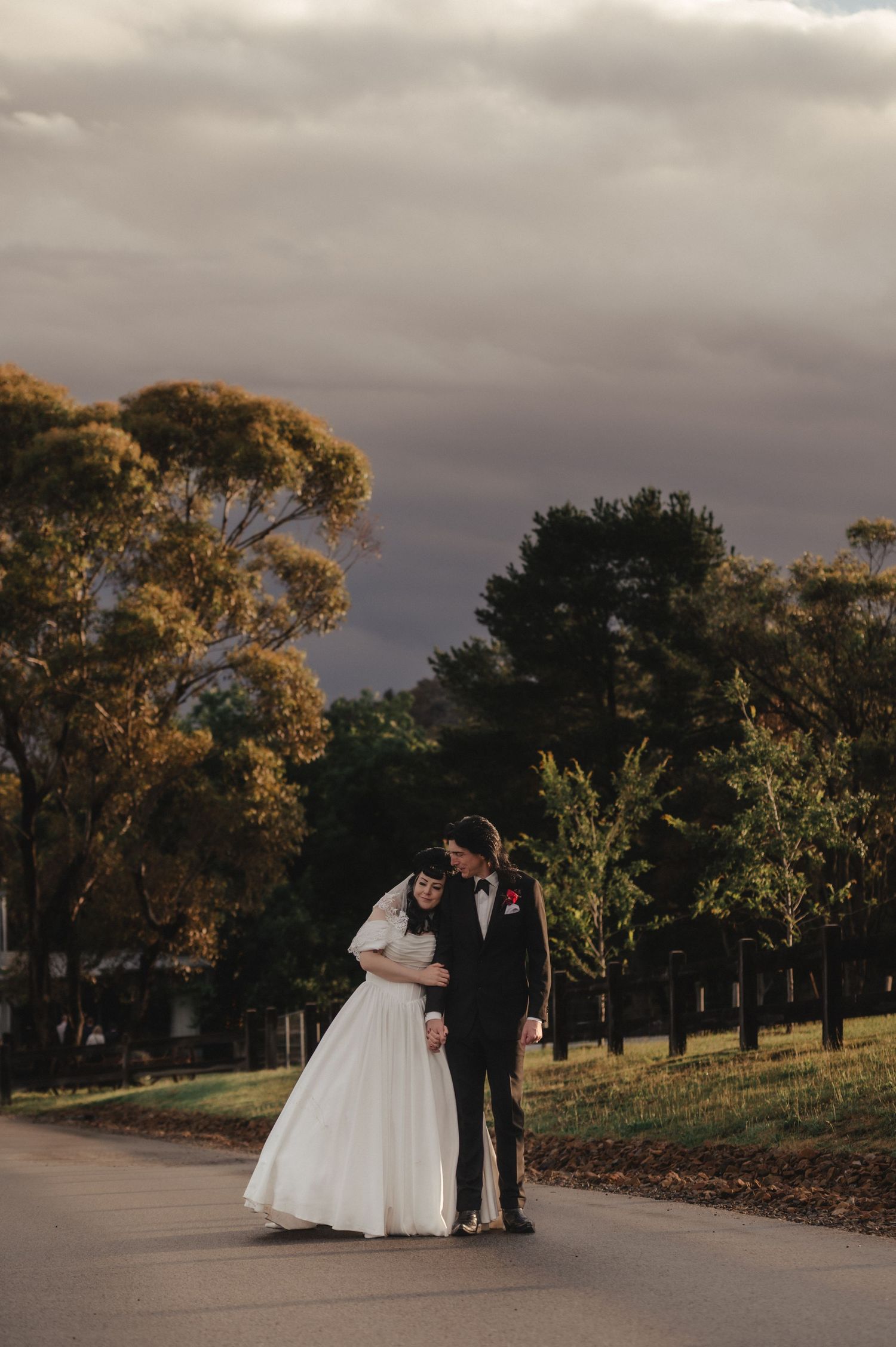 Bride and groom embrace on a path surrounded by golden trees under a dramatic cloudy sky.