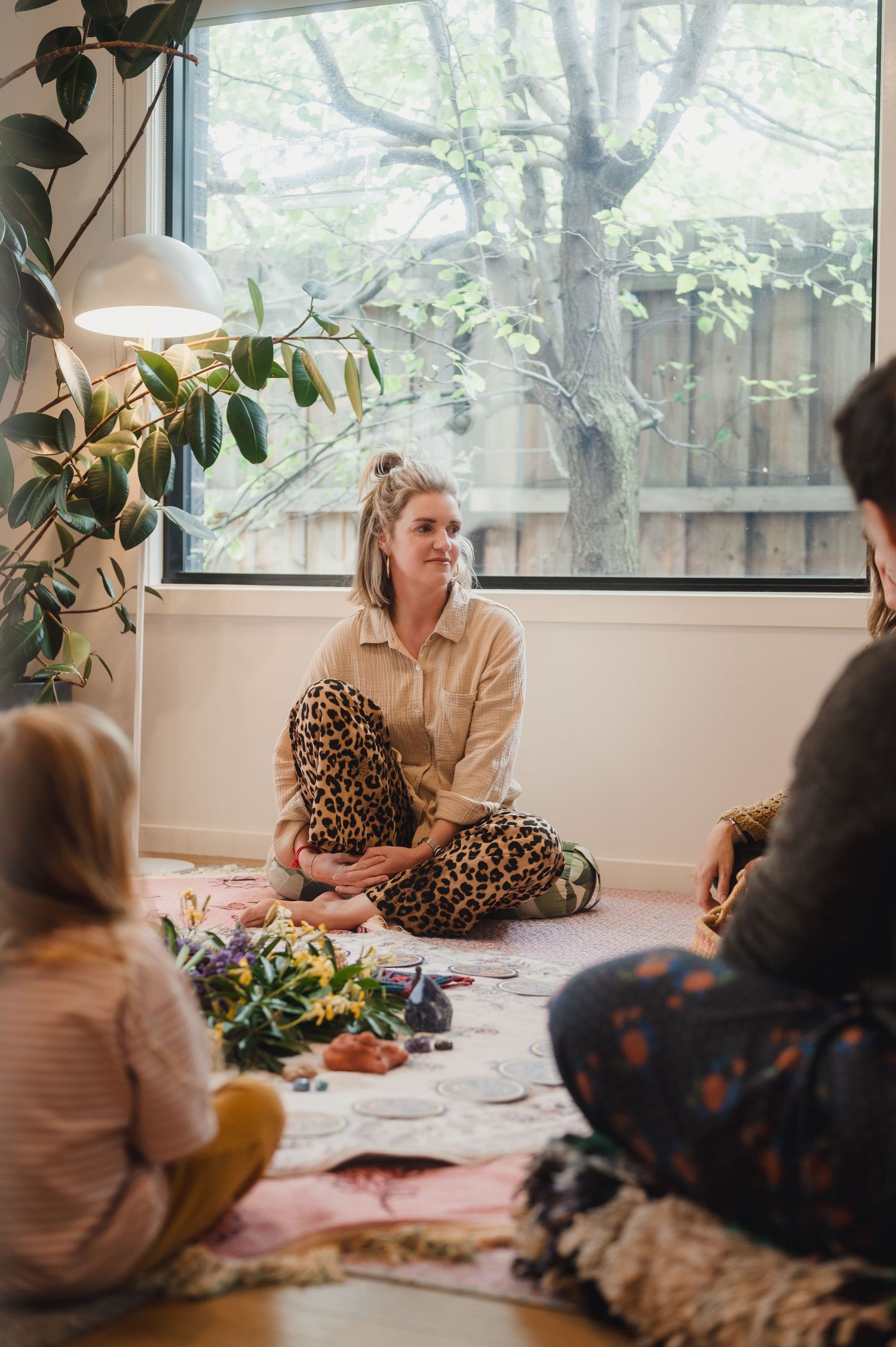 Woman in leopard print pants sits cross-legged on floor leading a small group wellness session indoors with plants and natural light.