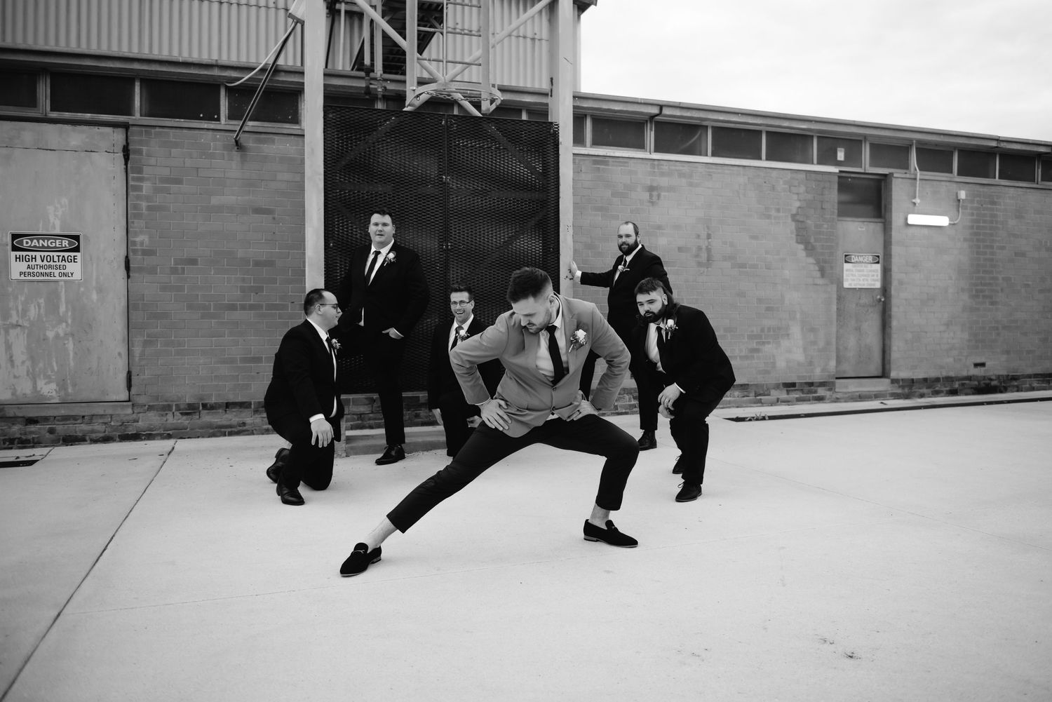 Wedding party jumps in unison against industrial building backdrop for fun group photo.