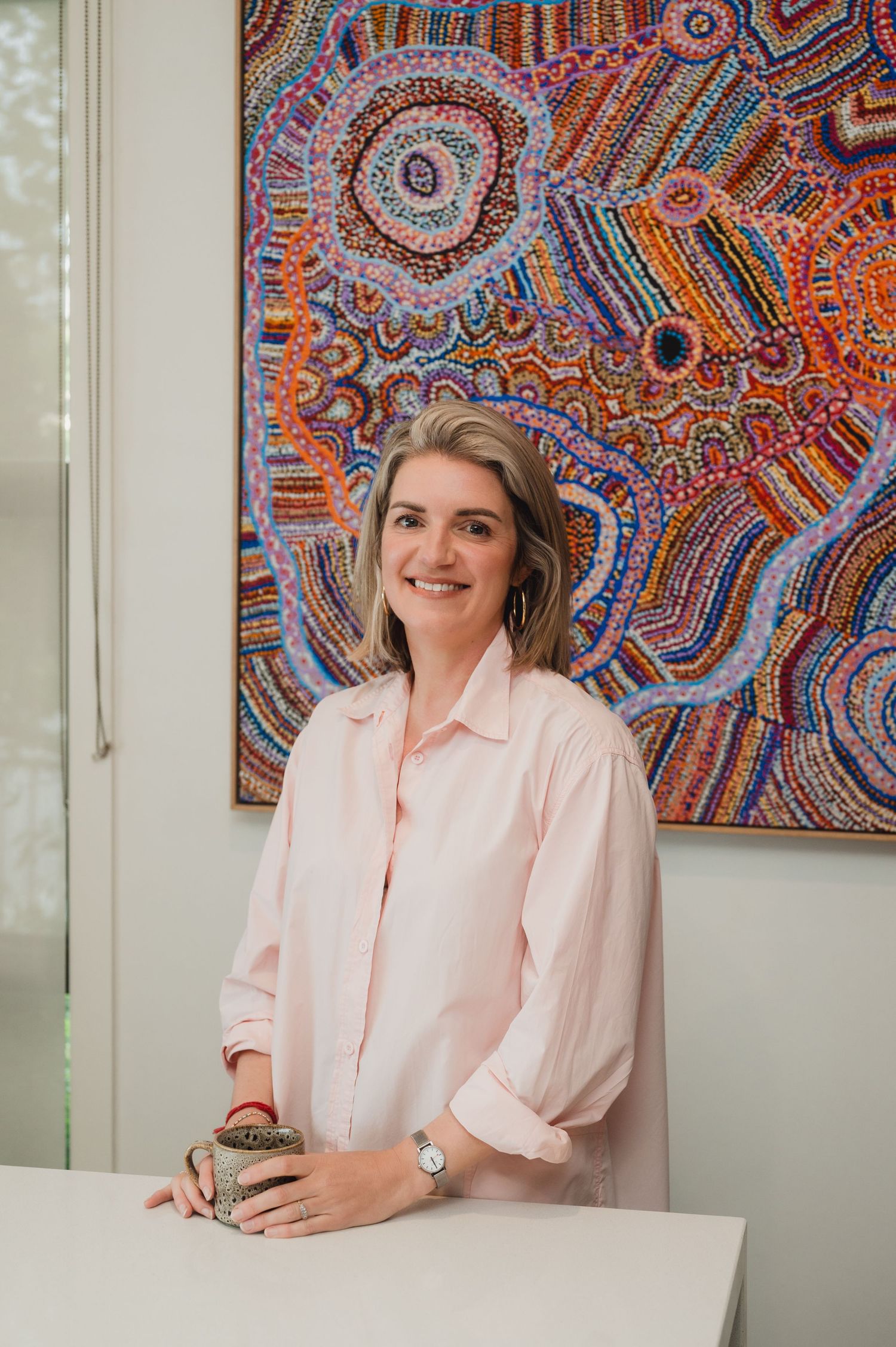 Woman in pink shirt smiling in front of colorful Aboriginal dot painting artwork.