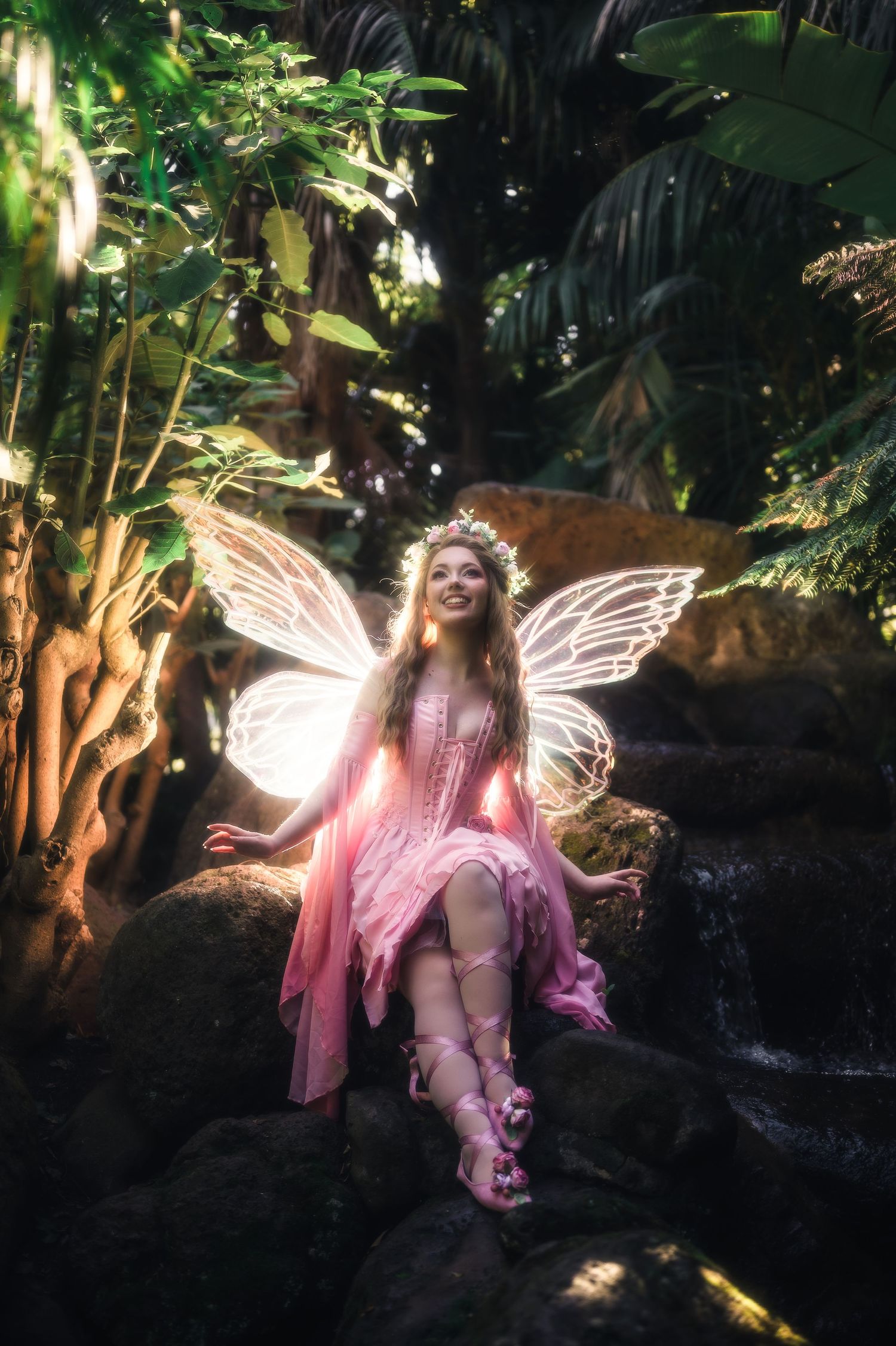 A woman in a pink fairy costume with glowing wings sits on rocks in a lush tropical garden.