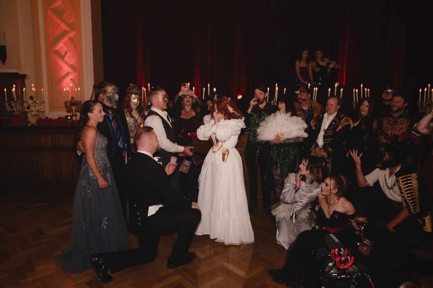 Costumed guests in elaborate period attire gather at a masquerade ball in a candlelit grand hall.