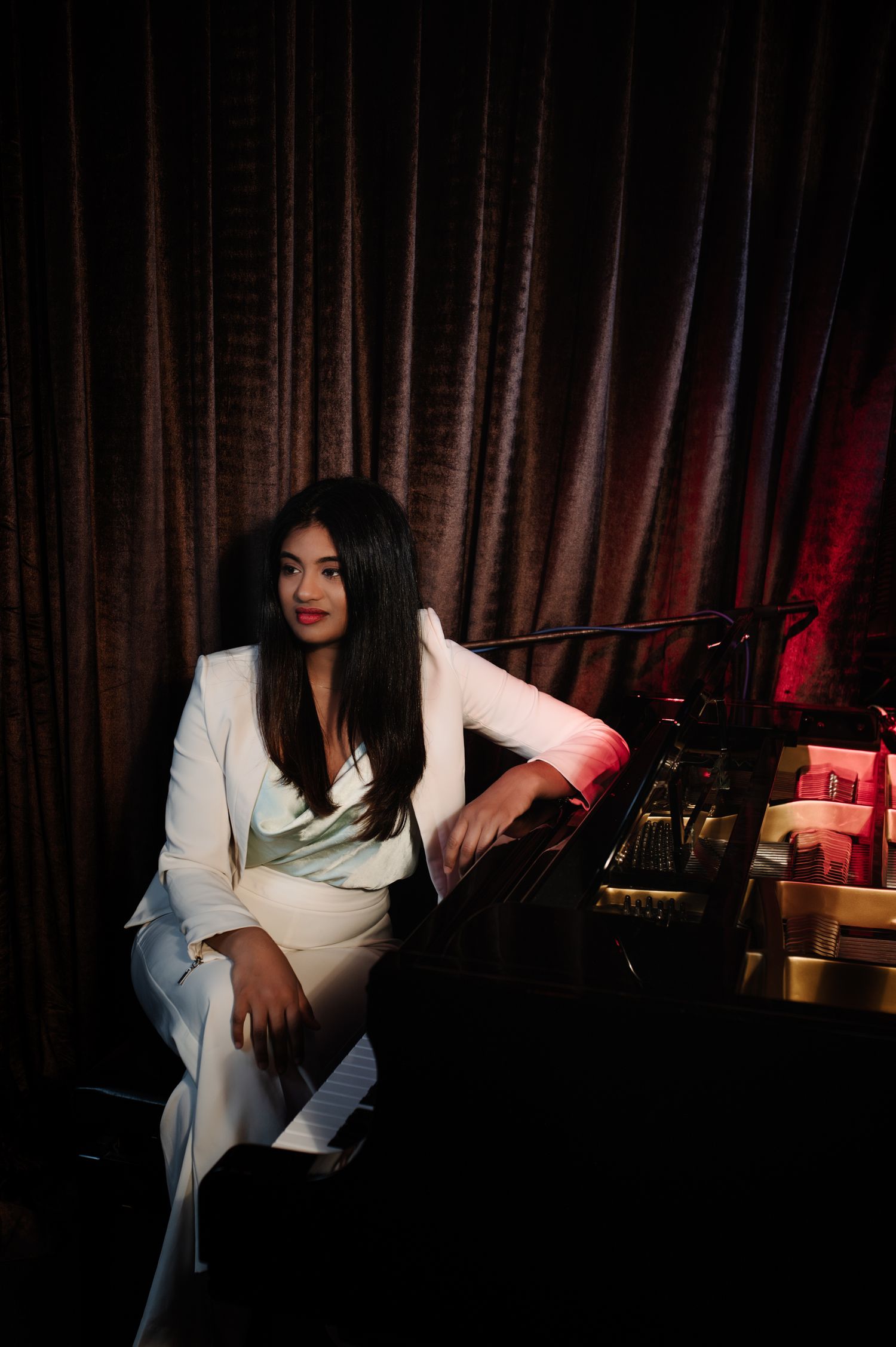 A musician in white attire performs expressively at a piano against dark curtains with red lighting.