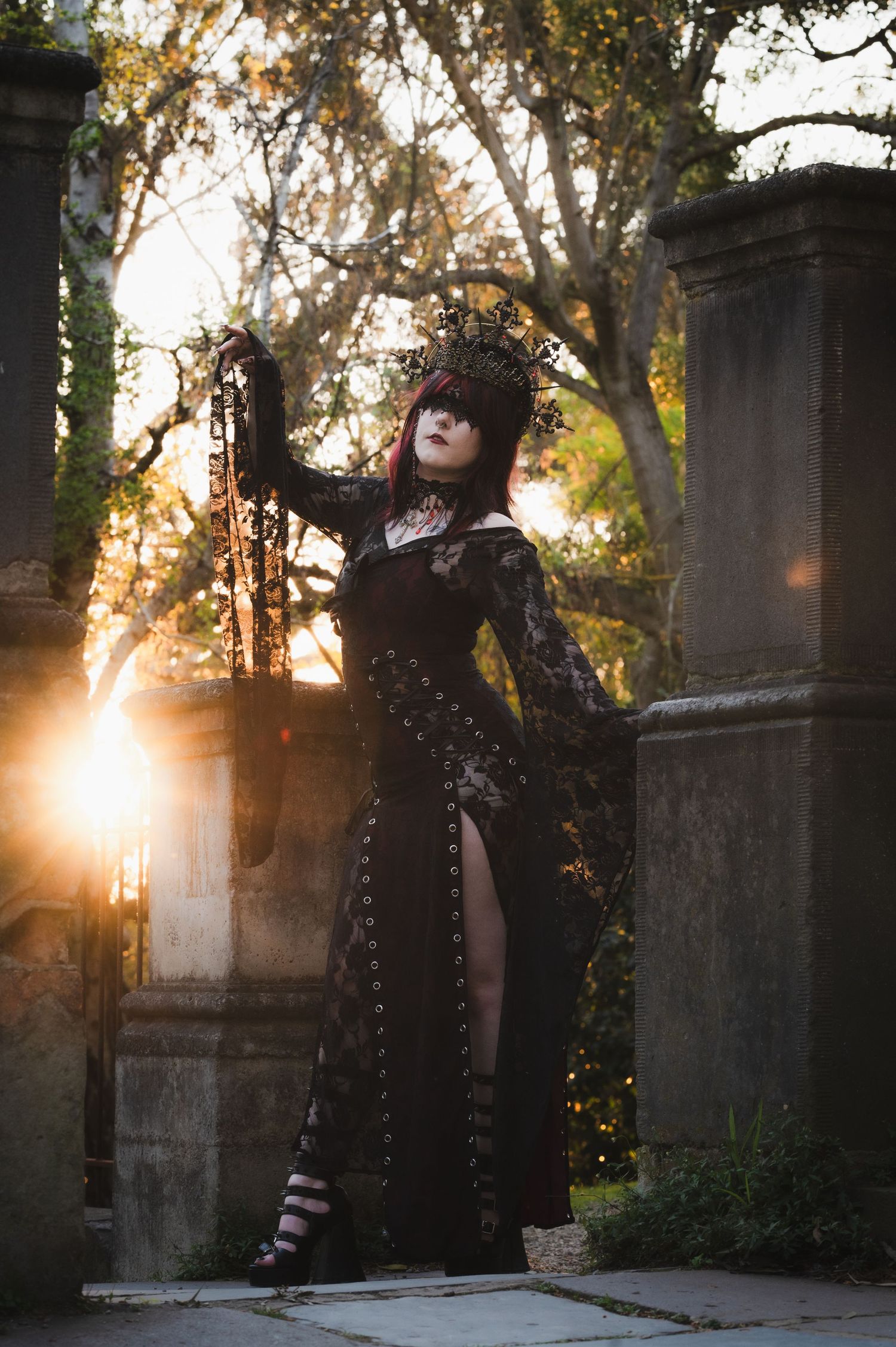 Gothic figure in black lace poses between stone pillars at sunset in a garden setting.