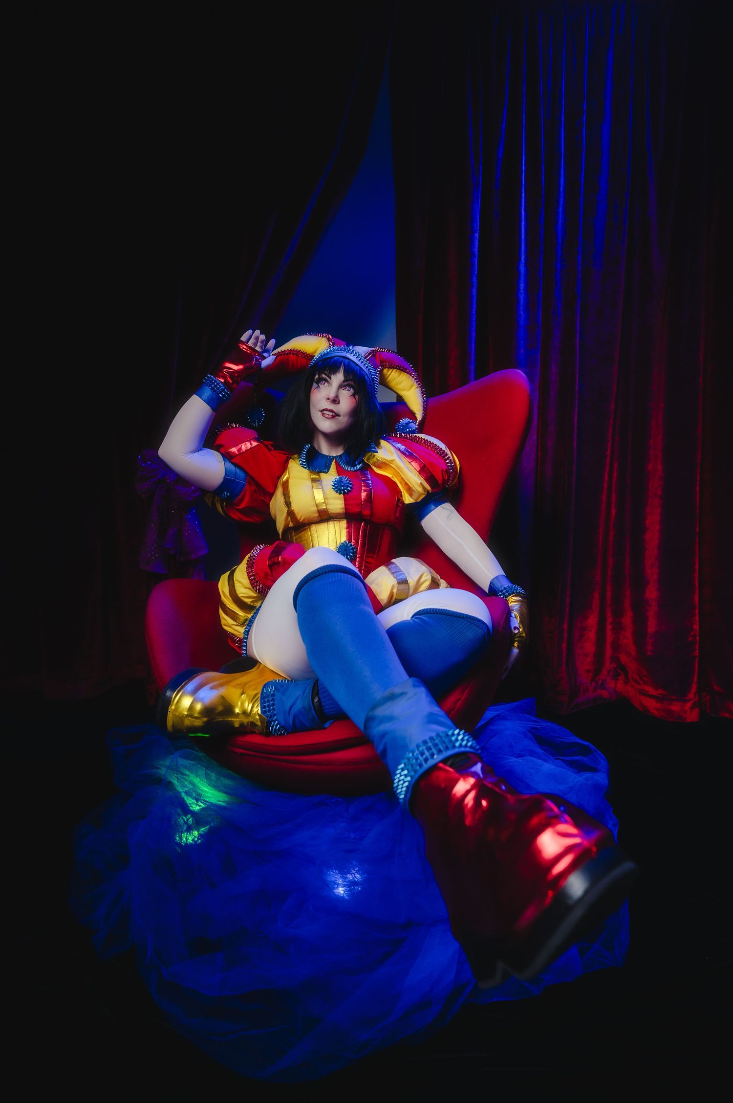 Cosplayer in colorful jester costume with blue thigh-highs and gold boots seated in red chair under dramatic stage lighting.