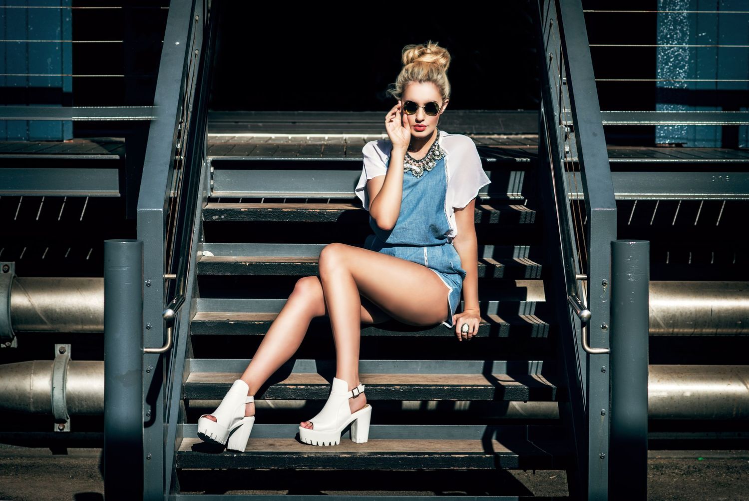 Stylish blonde woman in denim shorts and white heels sitting on metal stairs wearing sunglasses.