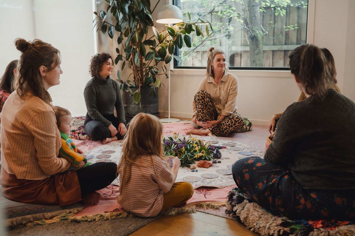 Women and children sit in a circle on colorful rugs during an indoor group meditation or mindfulness session.