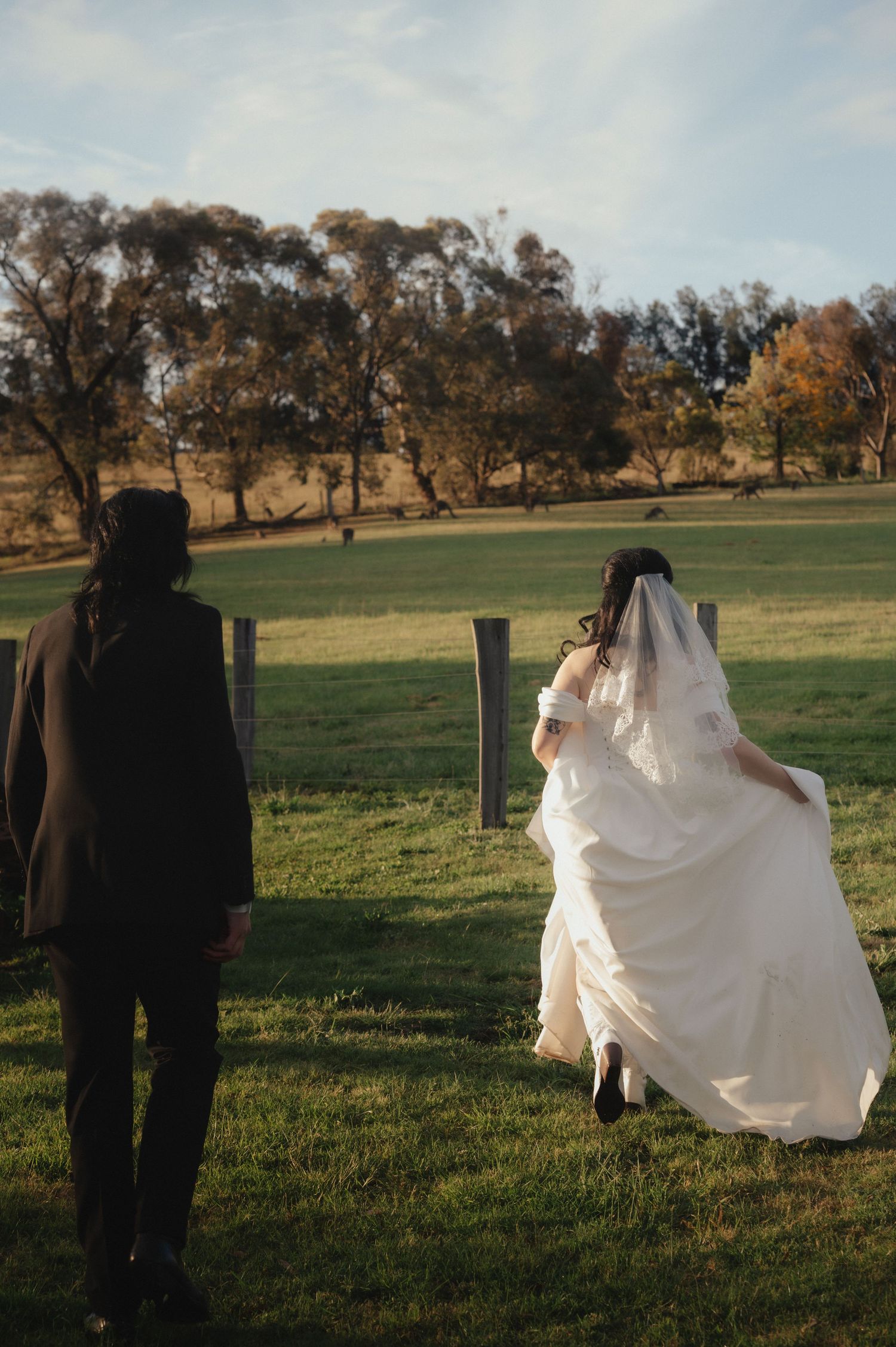 Bride and groom walking across a green field at a rural wedding venue, bride in white gown with veil flowing behind her.
