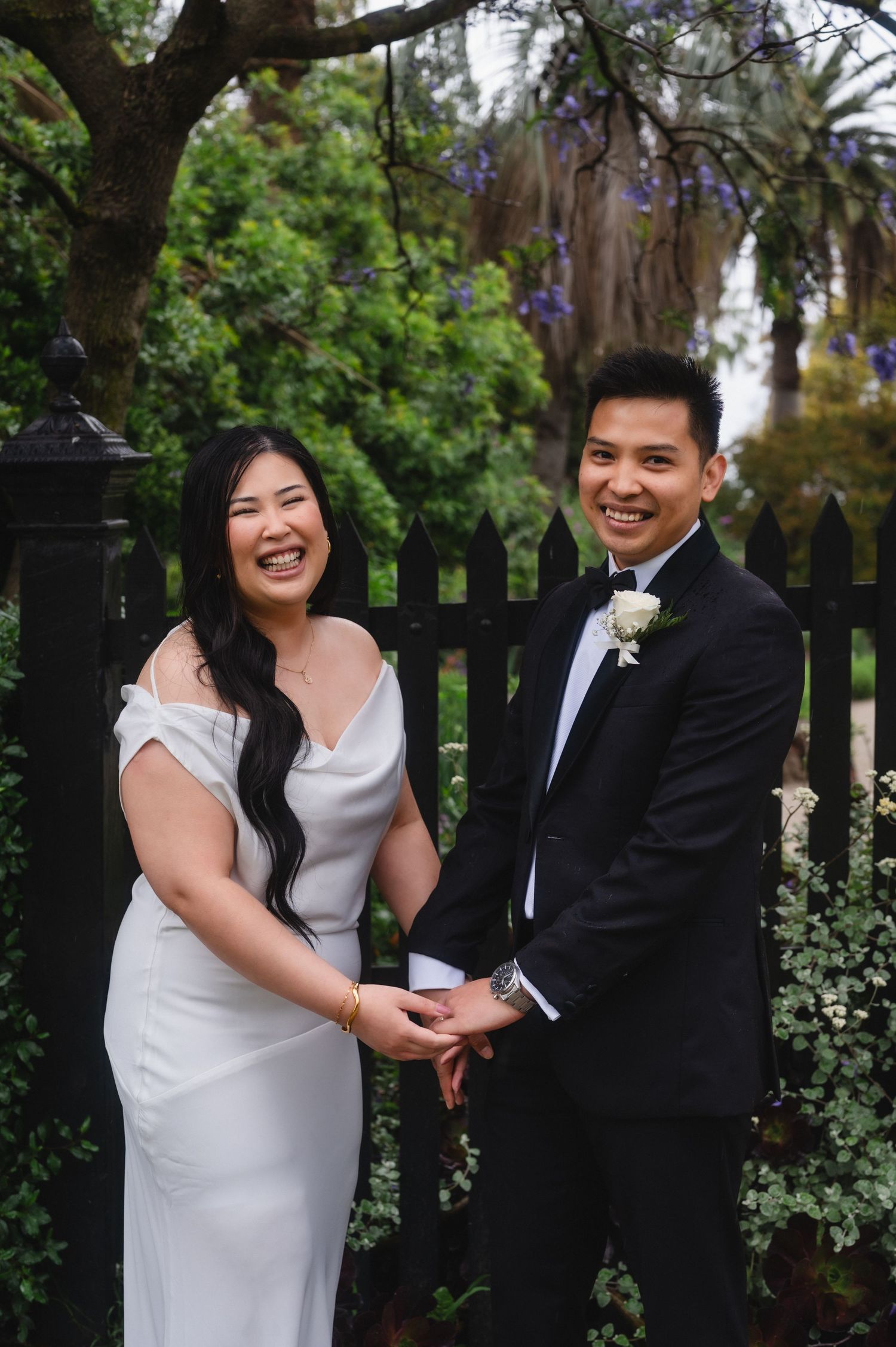 Smiling couple in wedding attire holding hands outdoors near black iron fence with lush green trees.