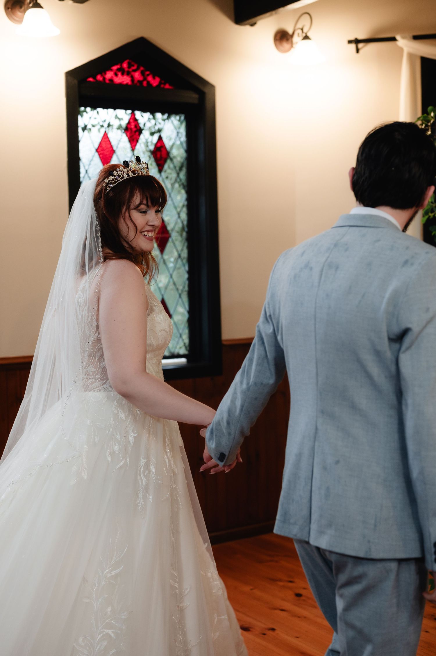 Wedding couple walks through indoor venue with diamond-patterned windows and wooden floors.