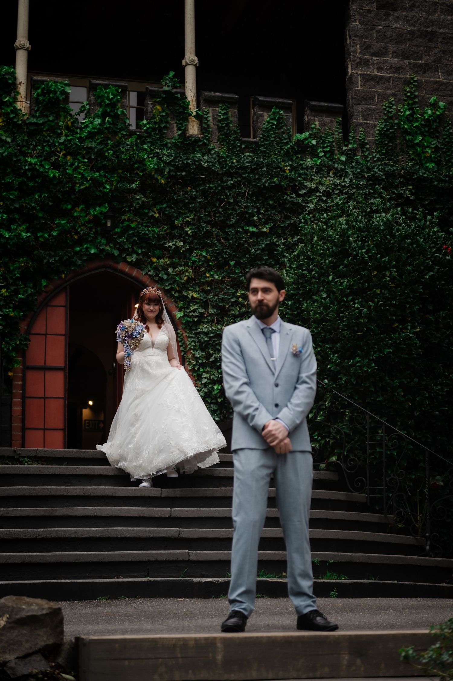 Bride in white lace gown and groom in grey suit pose playfully on outdoor steps.