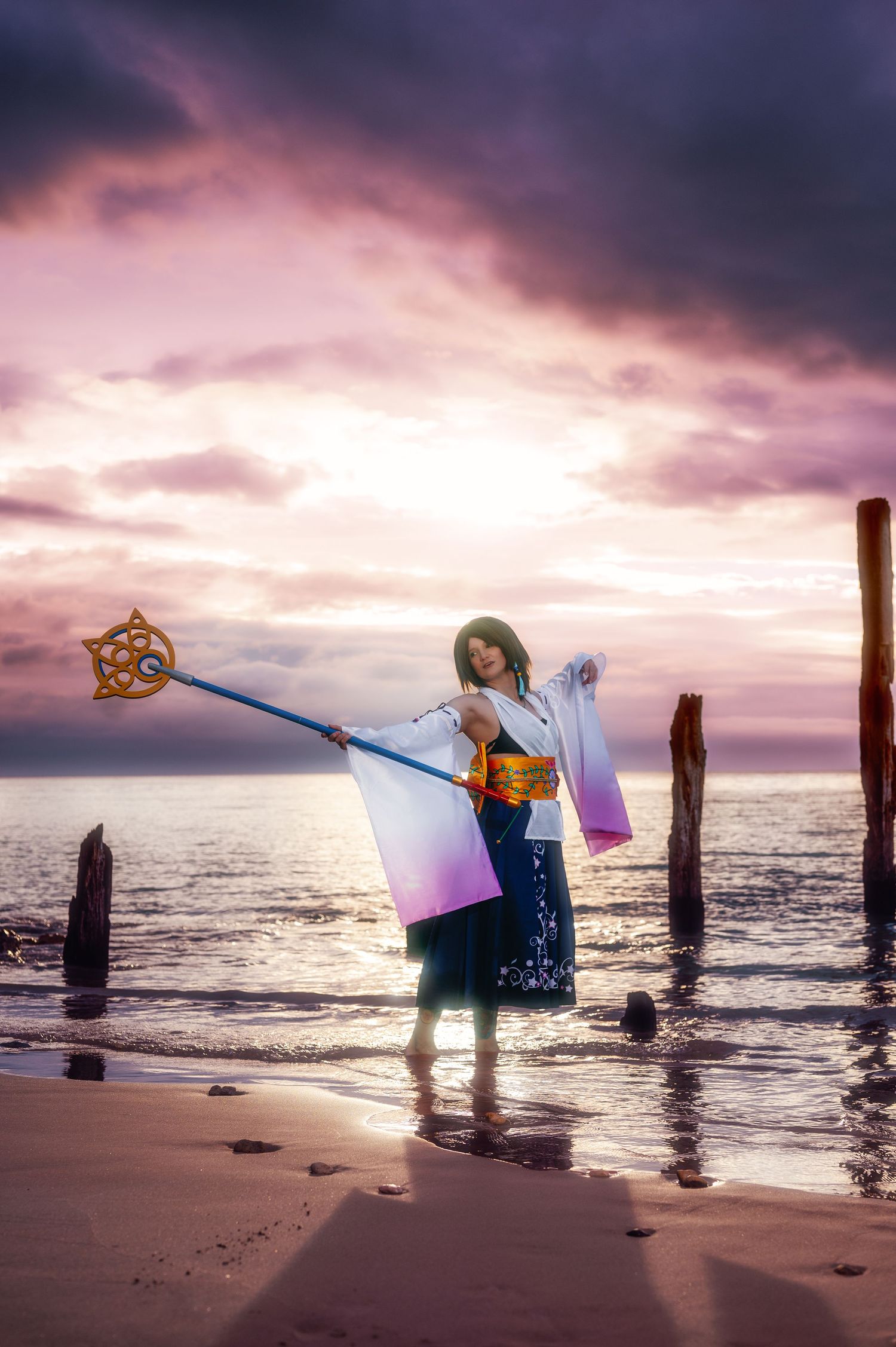 Cosplayer dressed as Yuna from Final Fantasy X poses on a beach at sunset holding a staff.