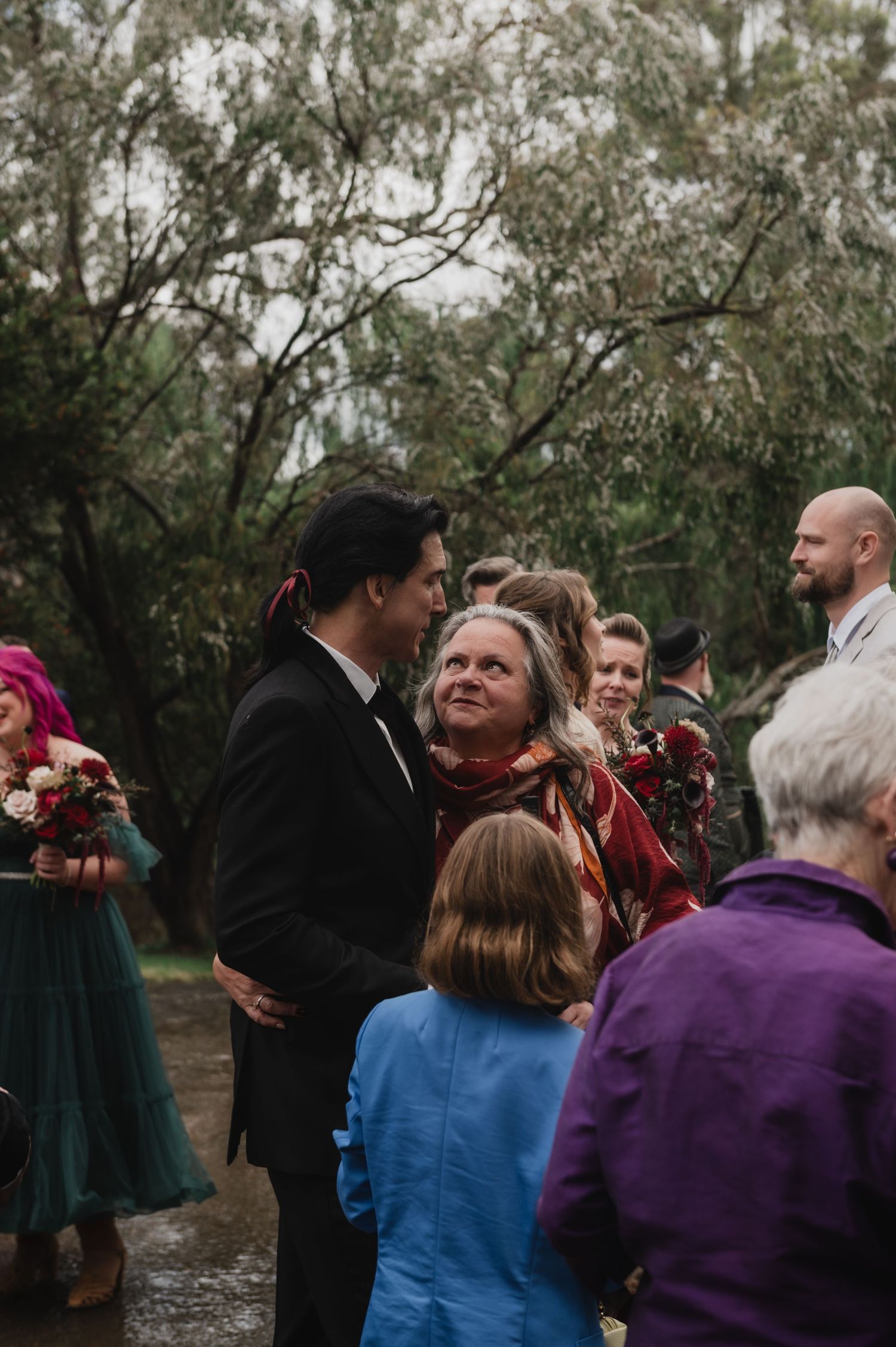 Guests mingle outdoors at a wedding reception surrounded by lush trees on a cloudy day.