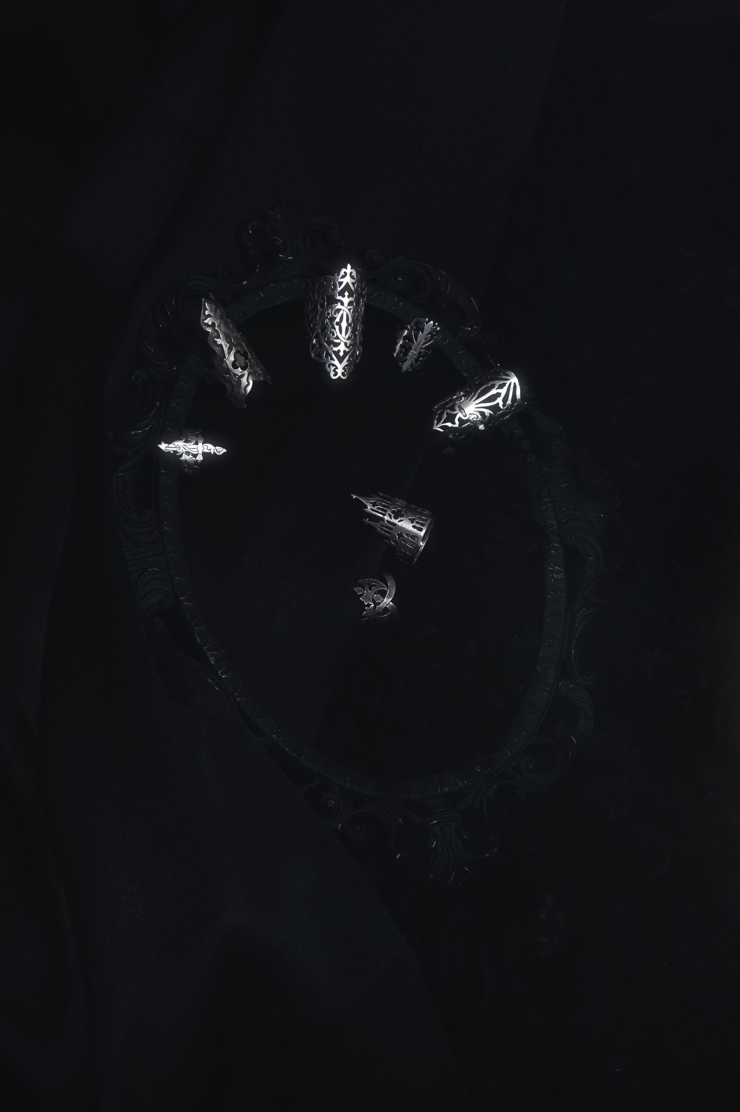 Dark abstract art featuring glowing silver ornate shapes arranged in a circular pattern on a black background.