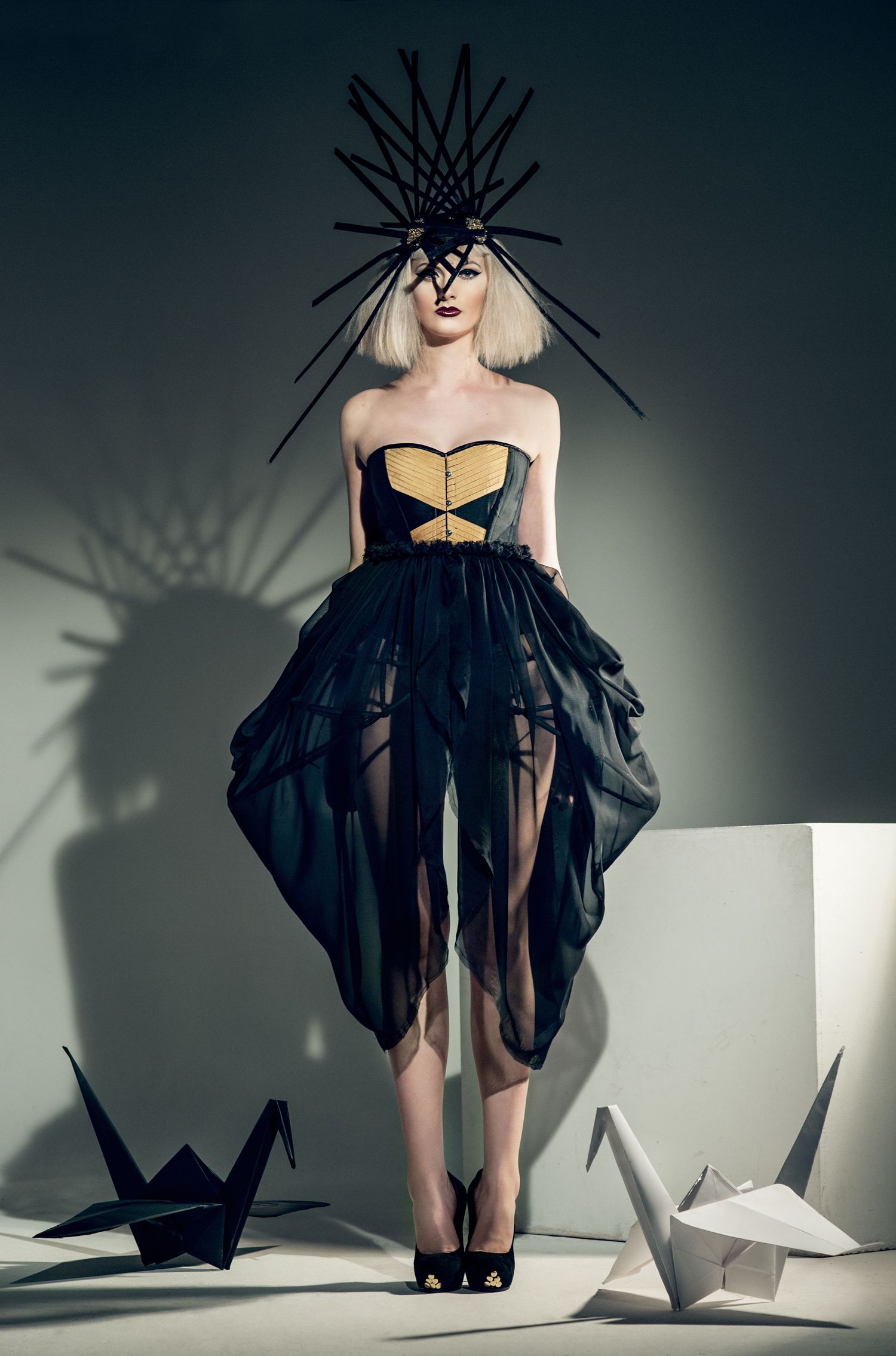 Model in black and gold strapless corset gown with sheer skirt, dramatic spiked headpiece, surrounded by origami cranes.