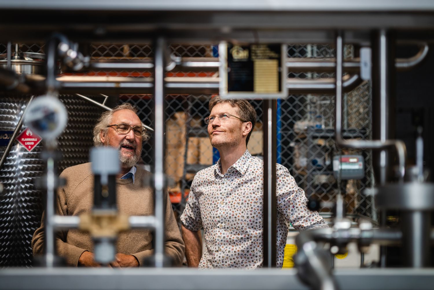 Two men conversing and smiling inside an industrial distillery surrounded by steel tanks and equipment.