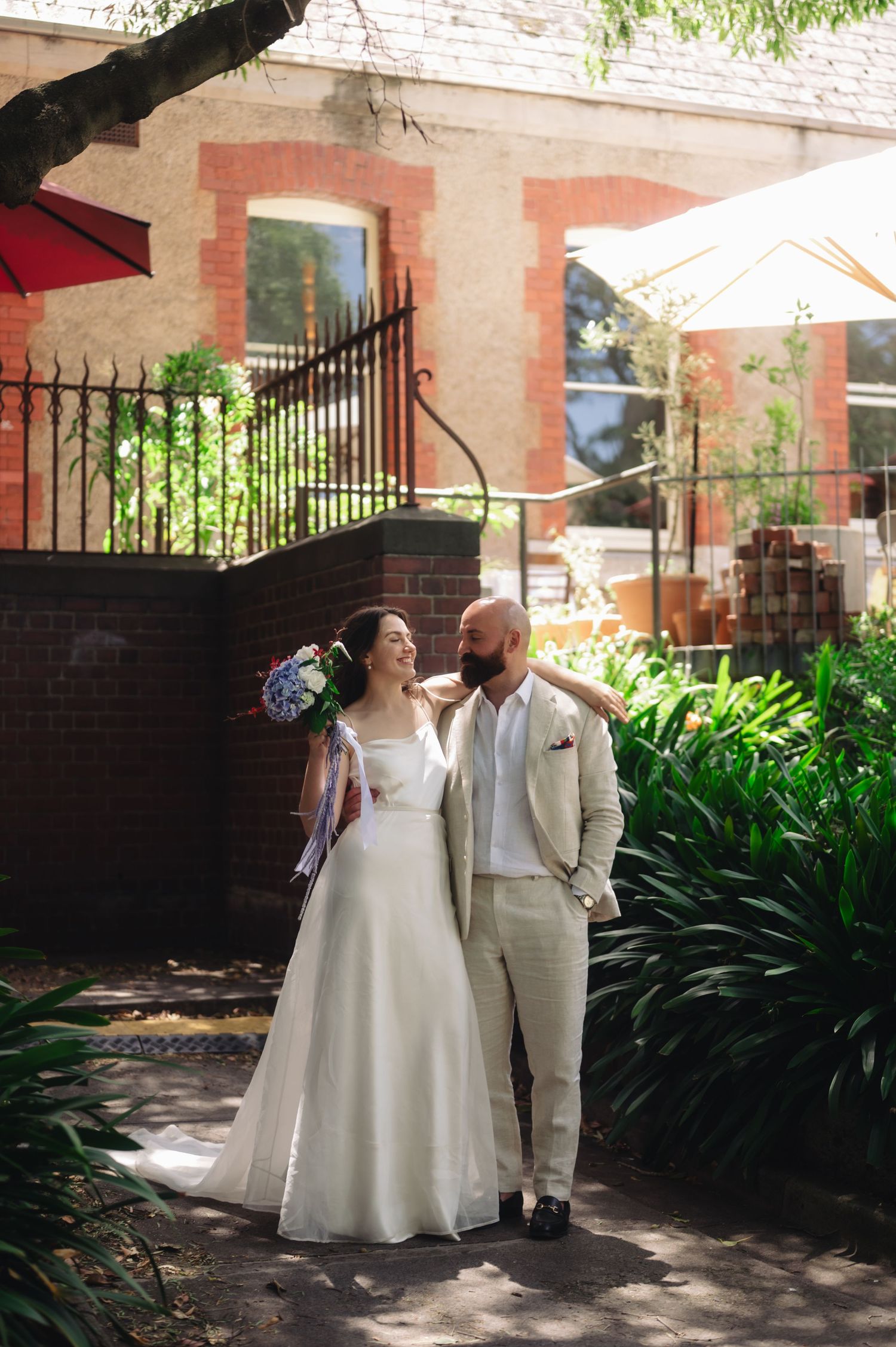 A bride and groom in wedding attire share a romantic moment in a sunlit garden courtyard beside a brick building.