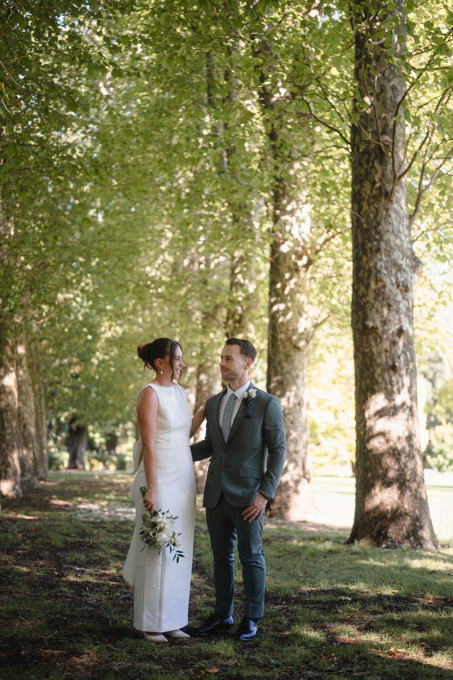 Bride and groom share a tender moment among tall trees in a sunlit park, she in white gown, he in olive suit.
