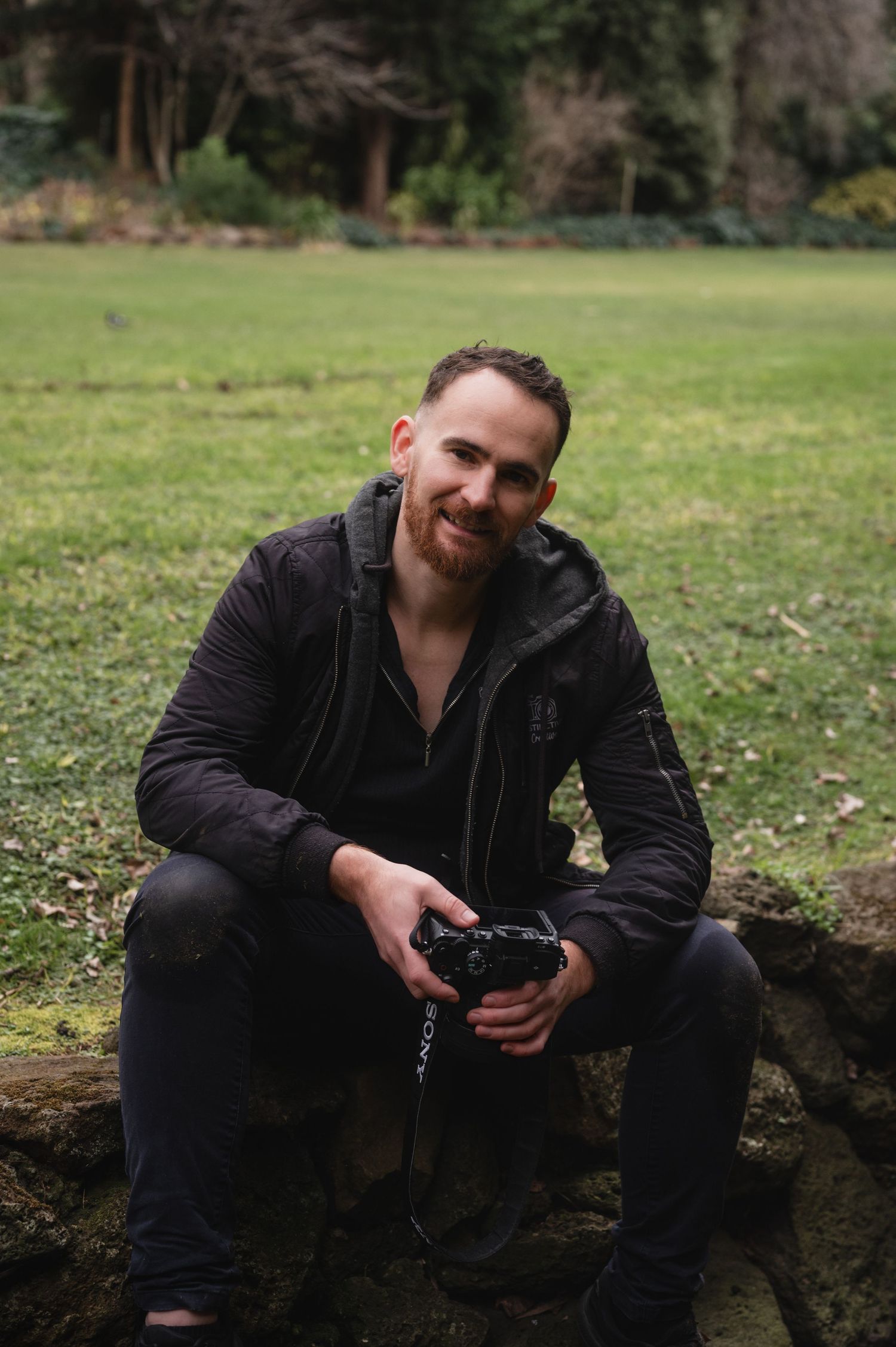 A smiling man with a red beard sits outdoors holding a camera, wearing a black jacket, with green grass in the background.