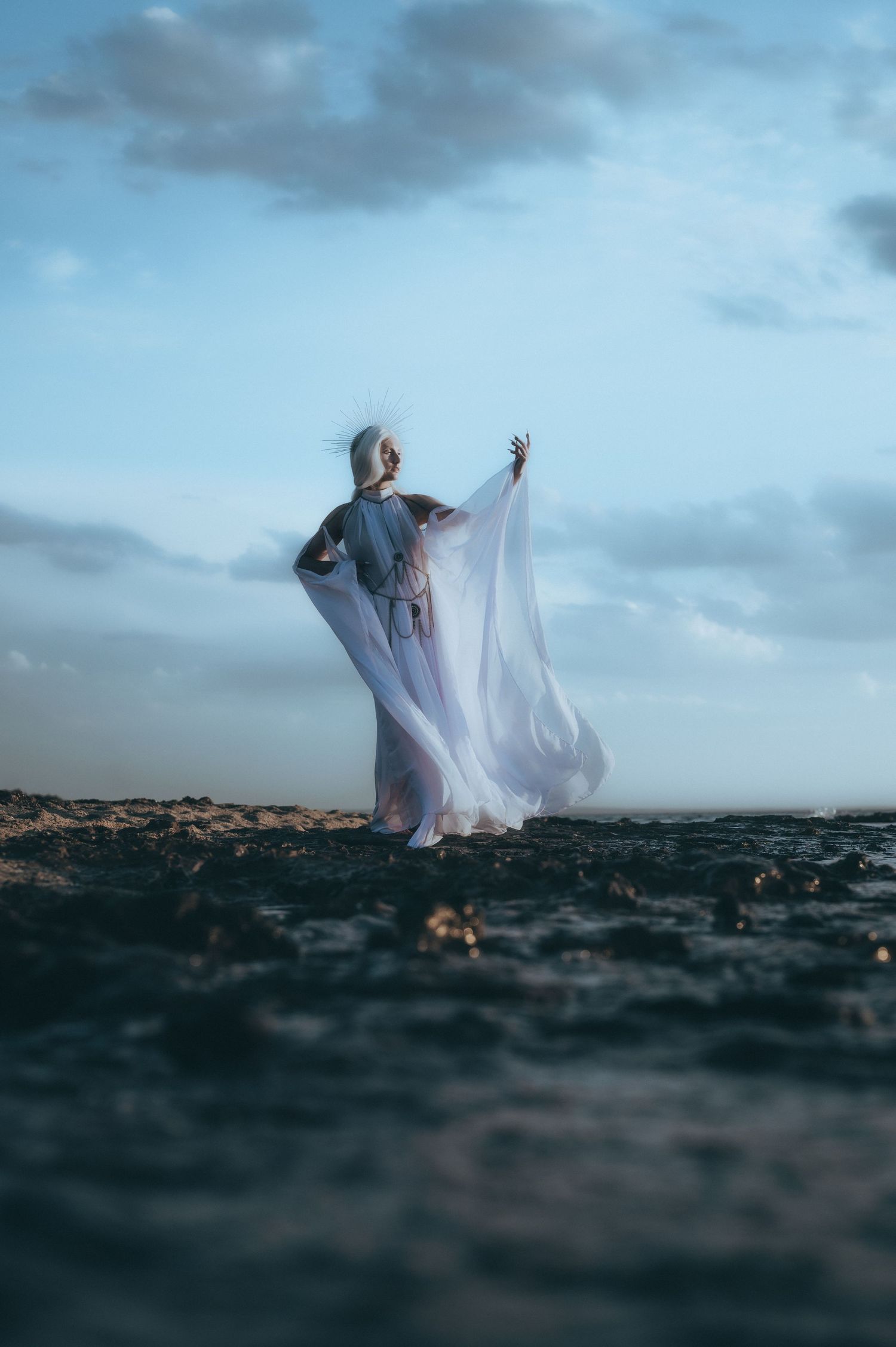 Ethereal figure in flowing white fabric dances on dark rocky terrain against moody cloudy sky.