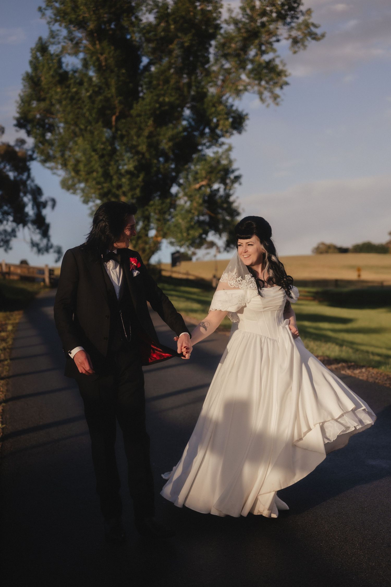 A romantic couple in wedding attire dances together on a path at sunset with trees and rolling hills in the background.