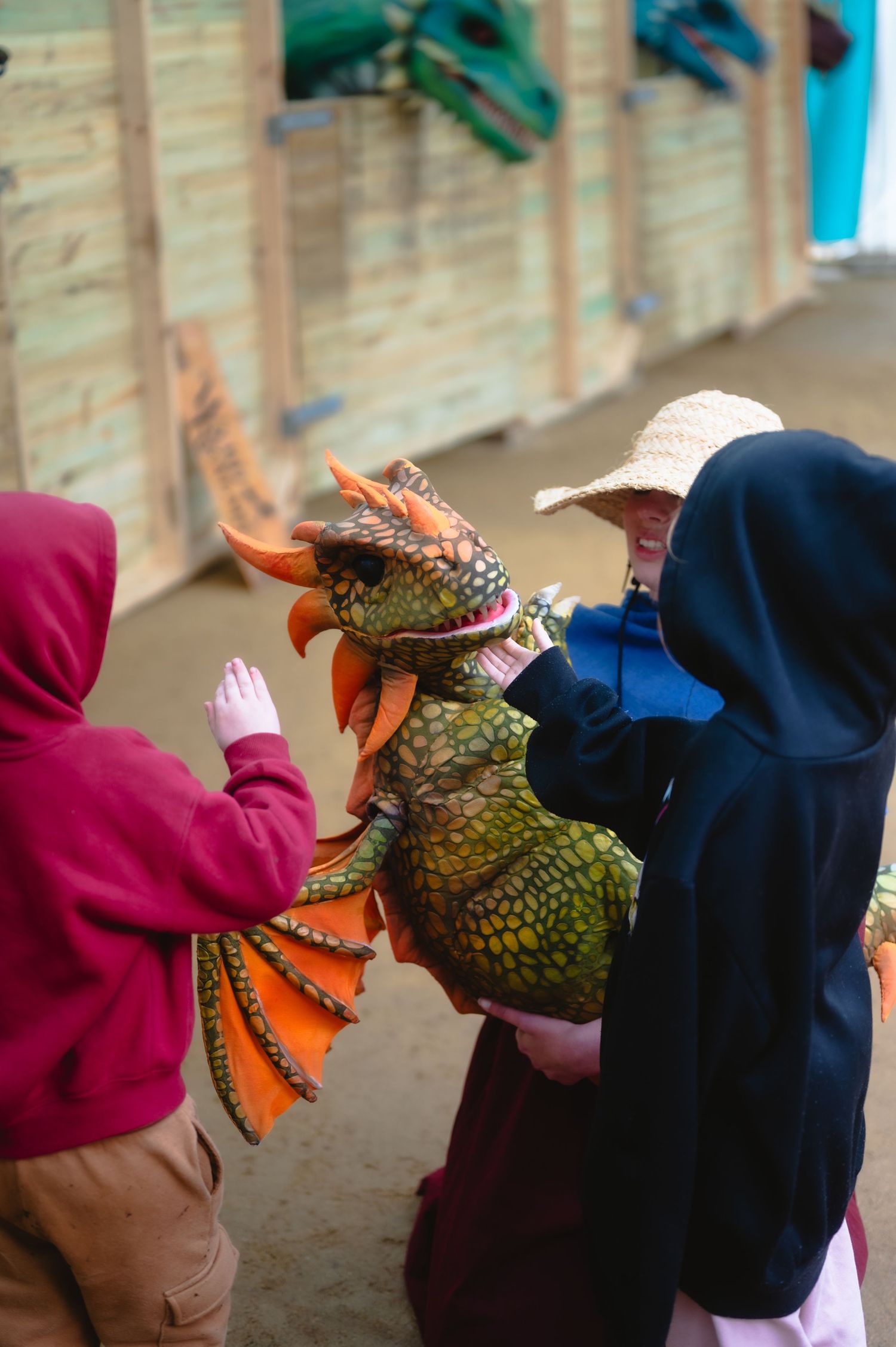 Children interact with a colorful dragon puppet at an outdoor medieval fair, with dragon decorations visible in the background.