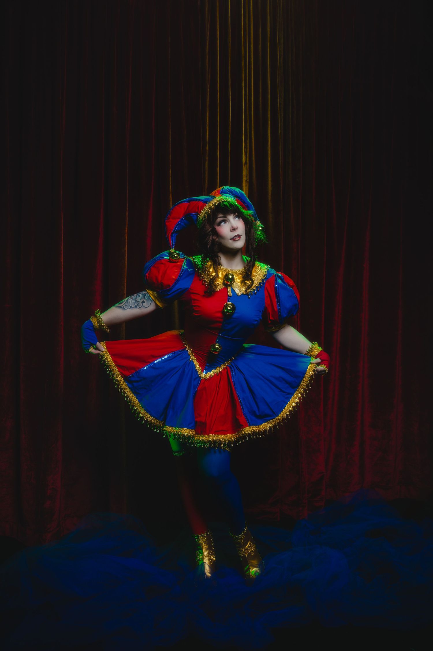 Performer in colorful jester costume with red, blue, and green dress poses dramatically on stage with red curtain backdrop.