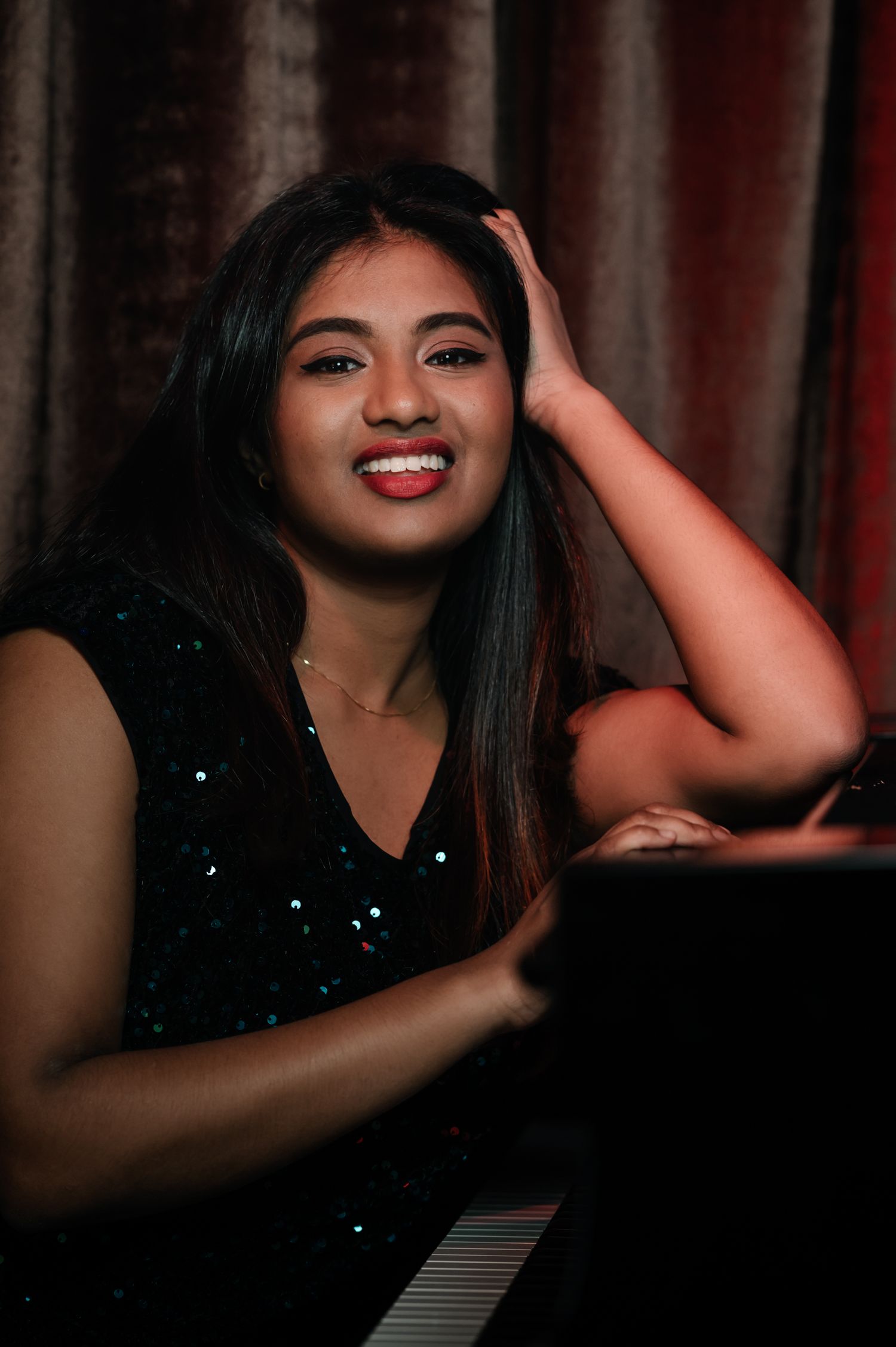 A performer in a sparkly black outfit poses playfully by a piano in moody lighting.