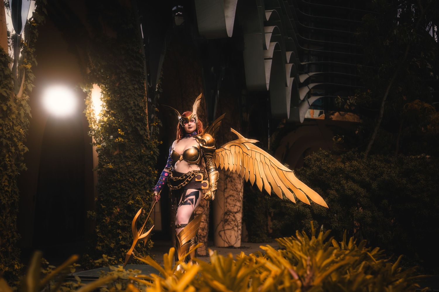Cosplayer dressed as a winged warrior goddess in golden armor poses dramatically in a lush garden at night.