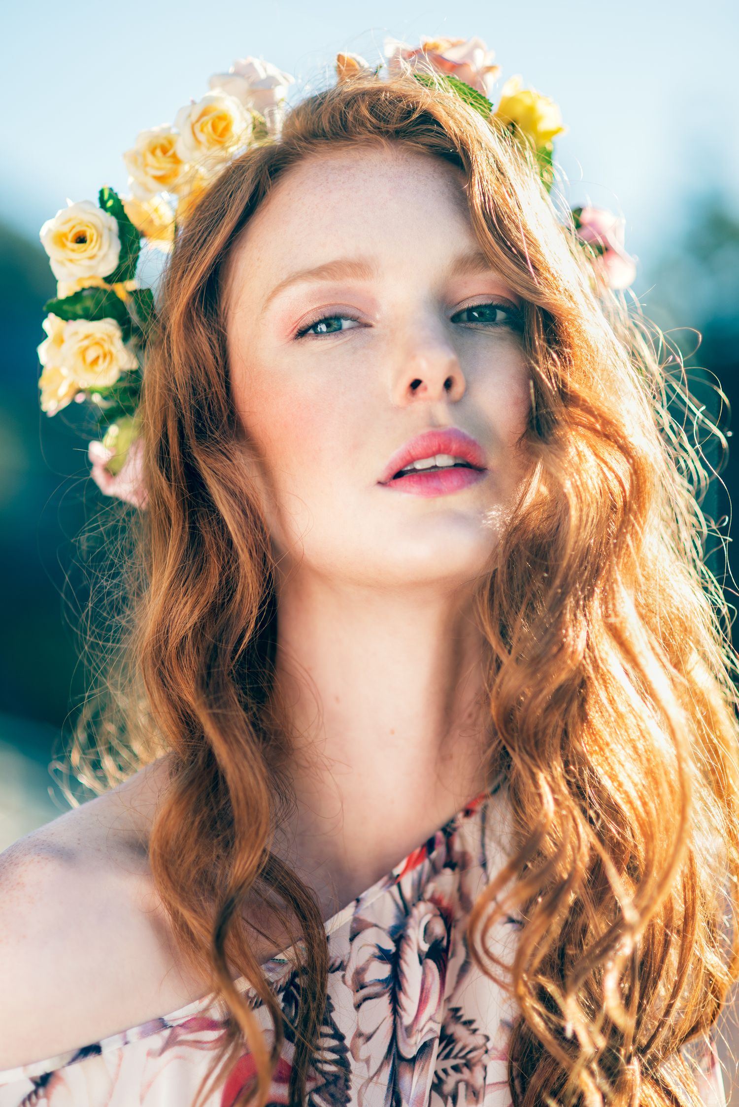 Young redhead woman with yellow flower crown and floral dress poses outdoors in soft sunlight.