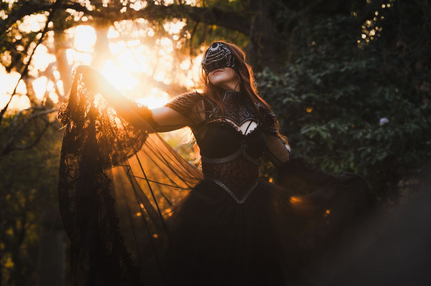 Woman in black fantasy costume with veil poses dramatically in golden sunset light among dark trees.