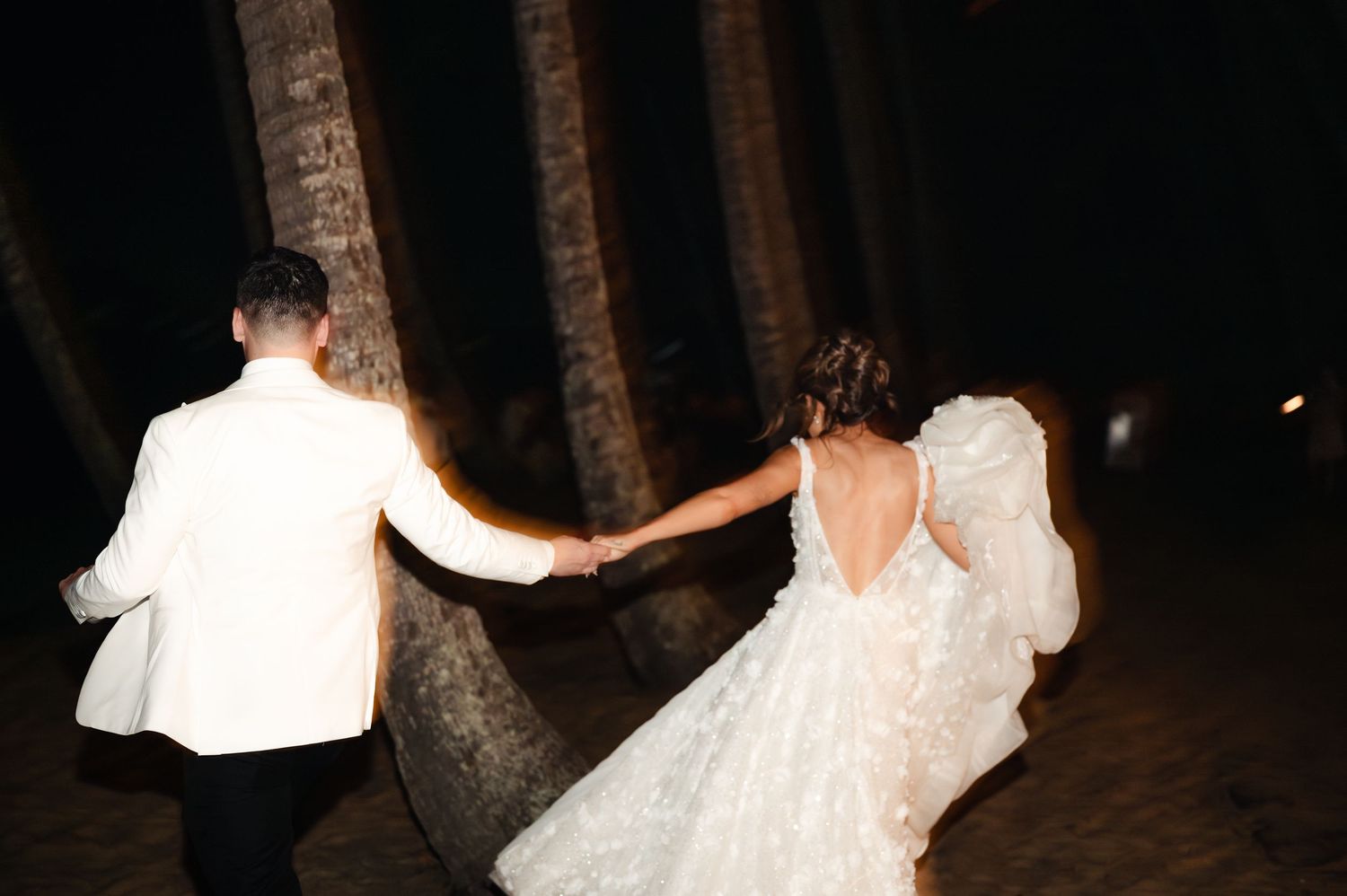 Couple dancing outdoors at night among palm trees at their wedding celebration.