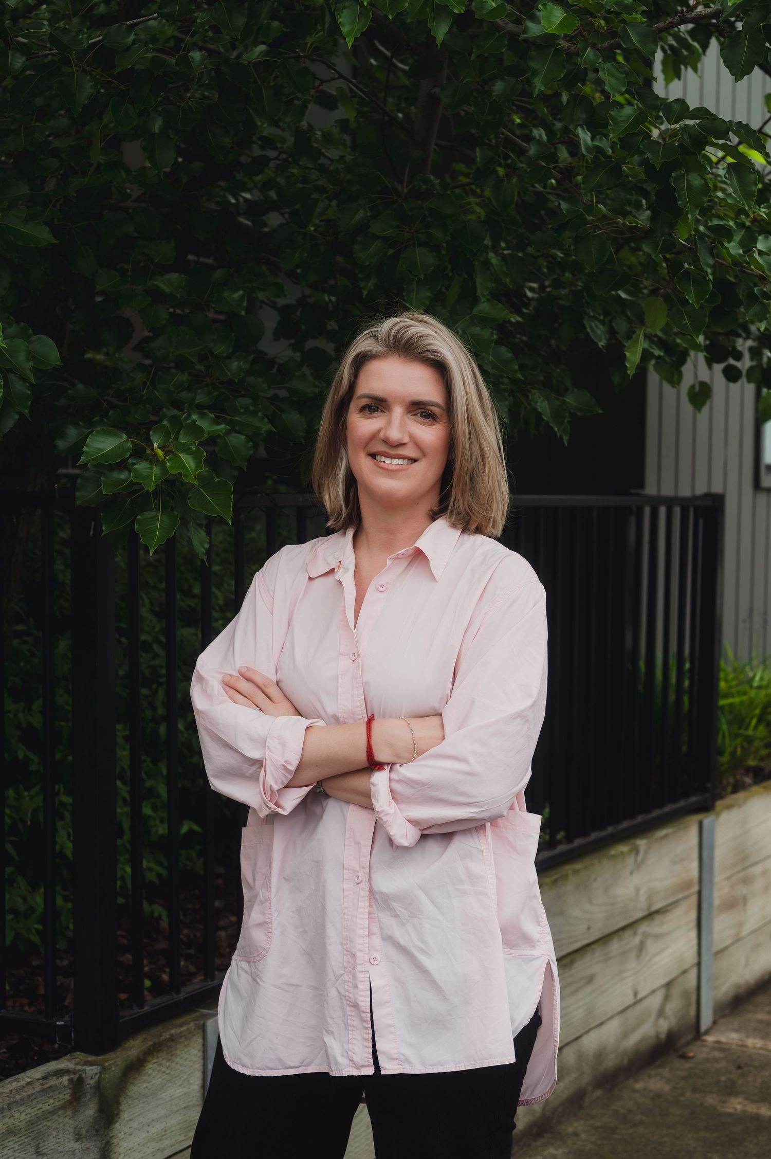 Smiling blonde woman in pink shirt standing with arms crossed outdoors near green trees and black iron fence.