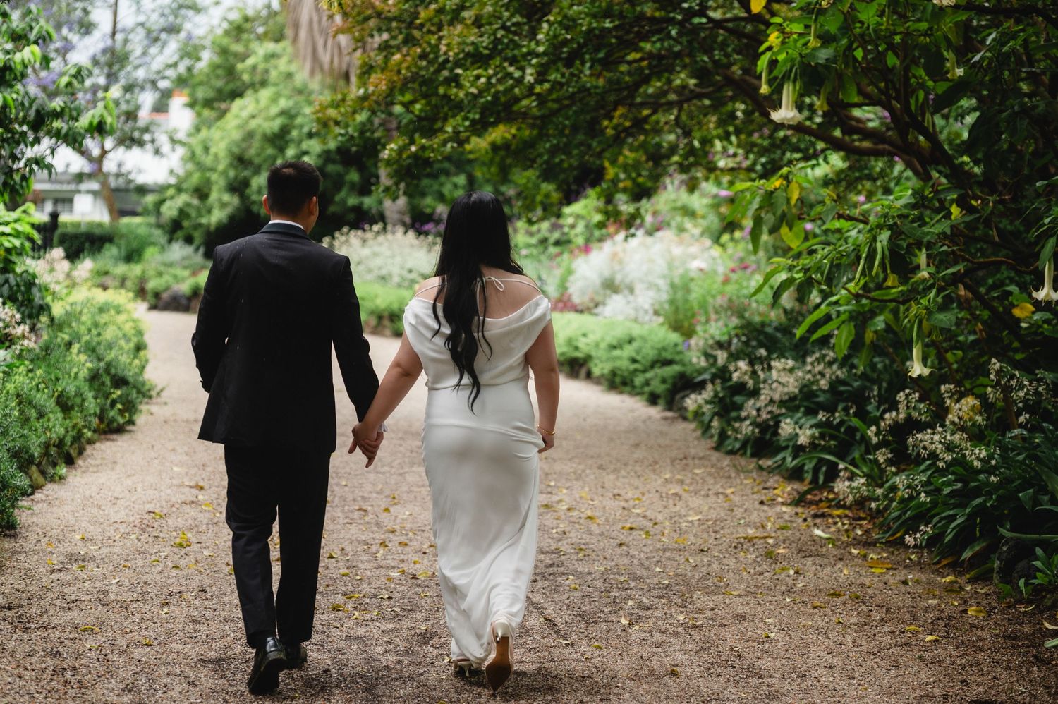 Bride and groom walking hand in hand along a garden path surrounded by lush greenery on their wedding day.